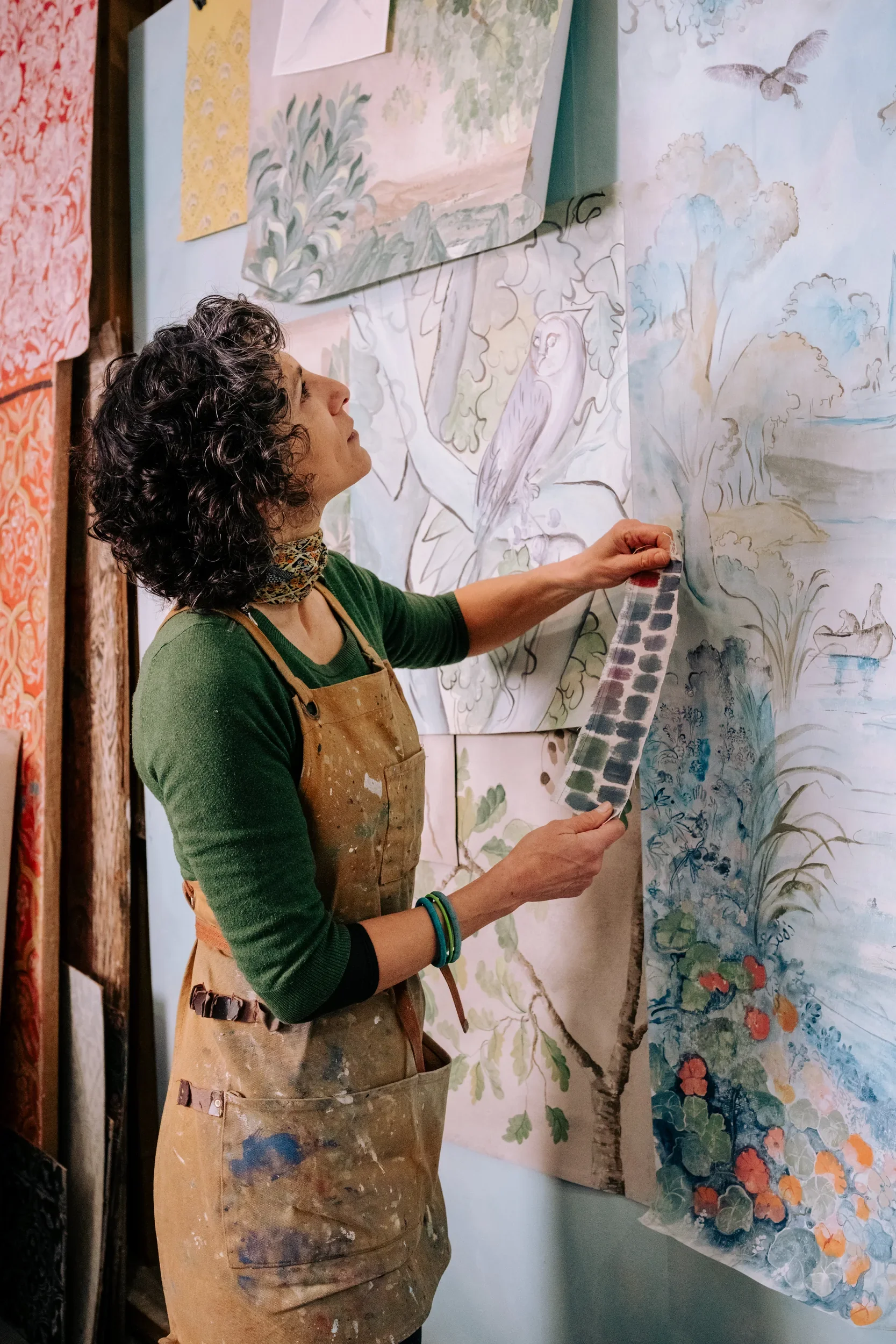 An artist wearing a green sweater, an apron, and jewelry is looking at a large watercolor painting of a landscape featuring a tree, flowers, and an owl, while holding a color palette.