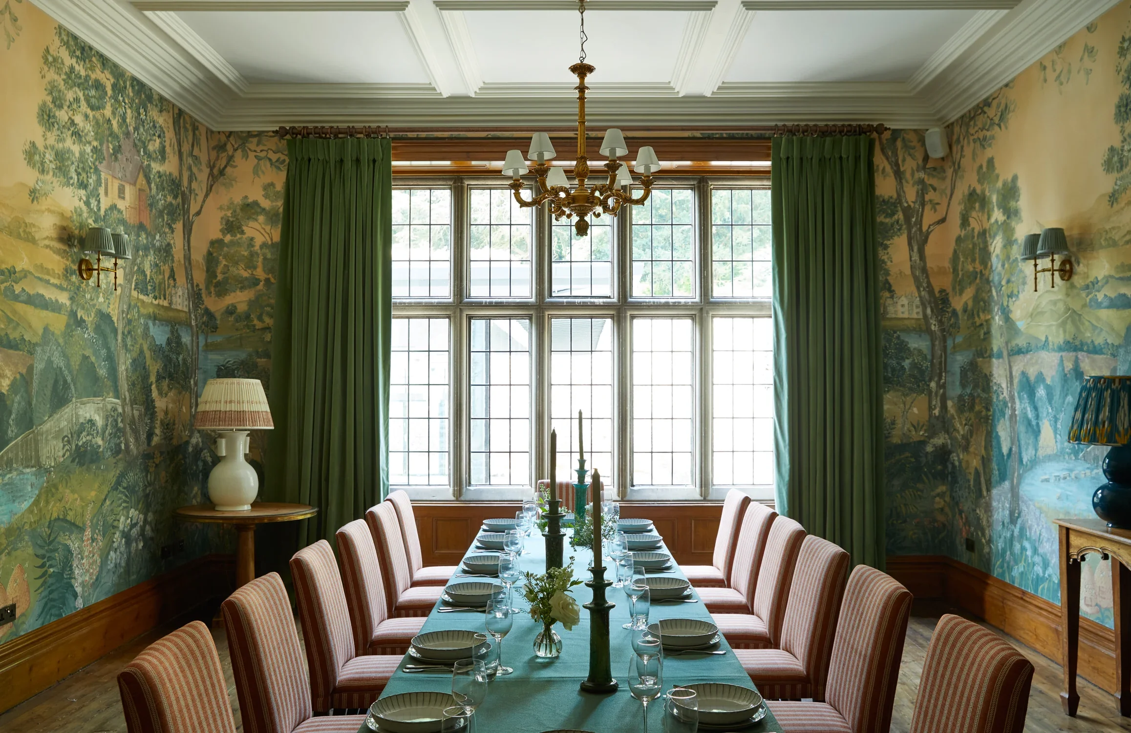 A formal dining room with large windows, green curtains, a chandelier, a long dining table set with plates, glasses, and napkins, surrounded by striped upholstered chairs, wallpaper with a landscape scene, and a side table with a table lamp.