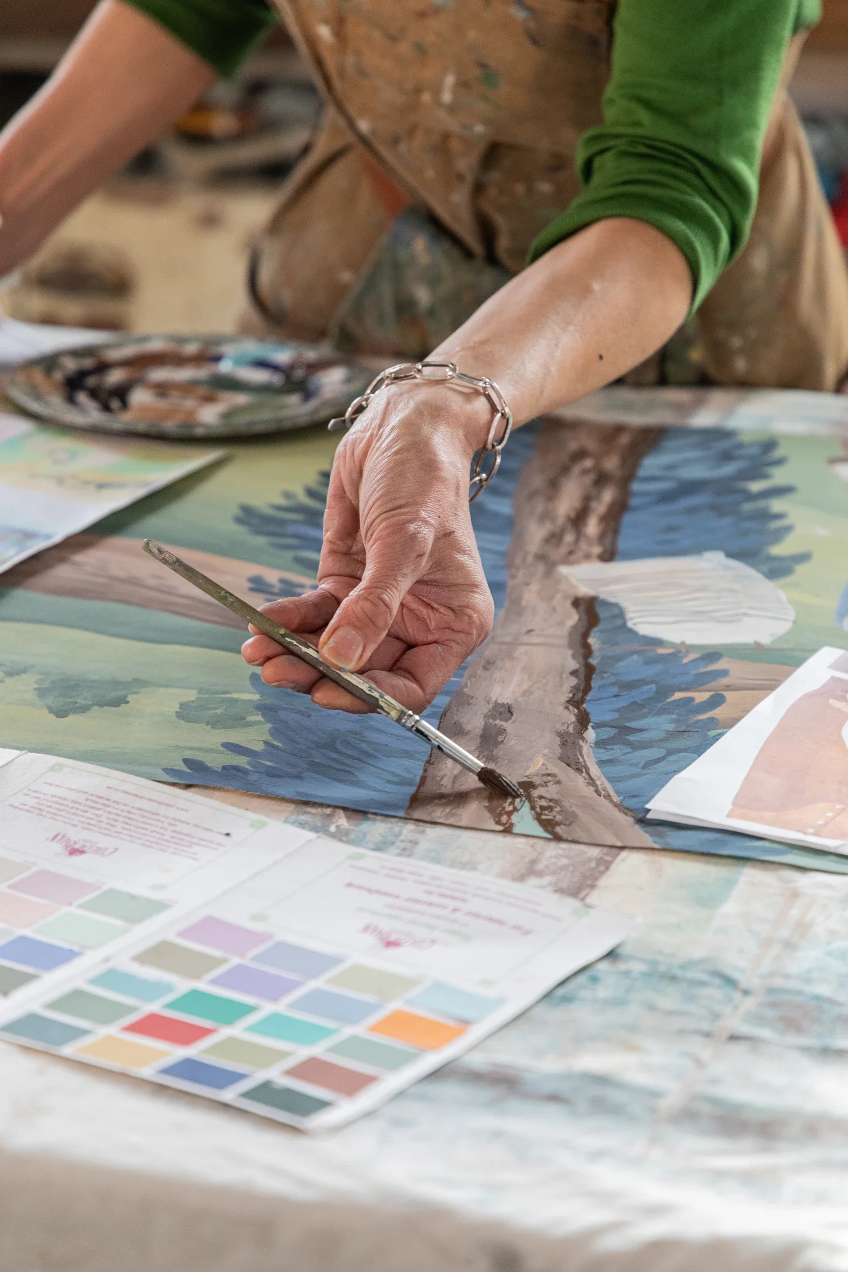 Photo shows a close up of Melissa White's hands holding a paintbrush hovering over a colour swatch and her own painting. She is in her Hastings studio.