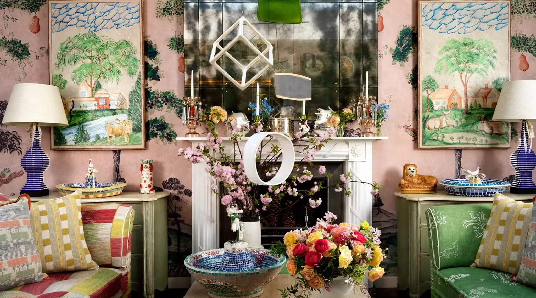 Living room decor with pink floral wallpaper, framed landscape paintings, two blue-striped lamps, a white fireplace mantel with a mirror above, pink cherry blossom branches, a large bouquet of flowers, ceramic animal figurines, and colorful pillows.