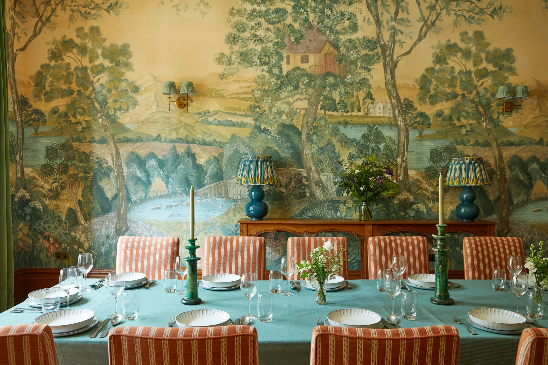Interior of a dining room with a large mural of a scenic landscape on the wall, featuring trees, a river, and buildings. The room has a long table set with plates, glasses, and cutlery, with a centerpiece of flowers and blue tablecloth. There are two blue lamps and two green candleholders on the table.