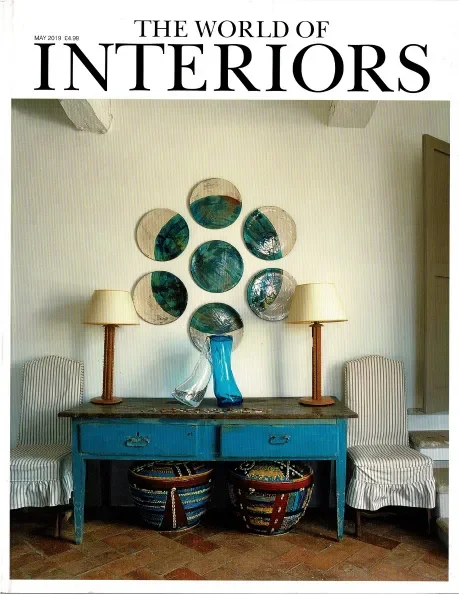 Cover of 'The World of Interiors' magazine featuring an interior decor scene with a blue console table, striped armchairs, lamps, colorful woven baskets underneath, and wall mirrors arranged in a circular pattern.