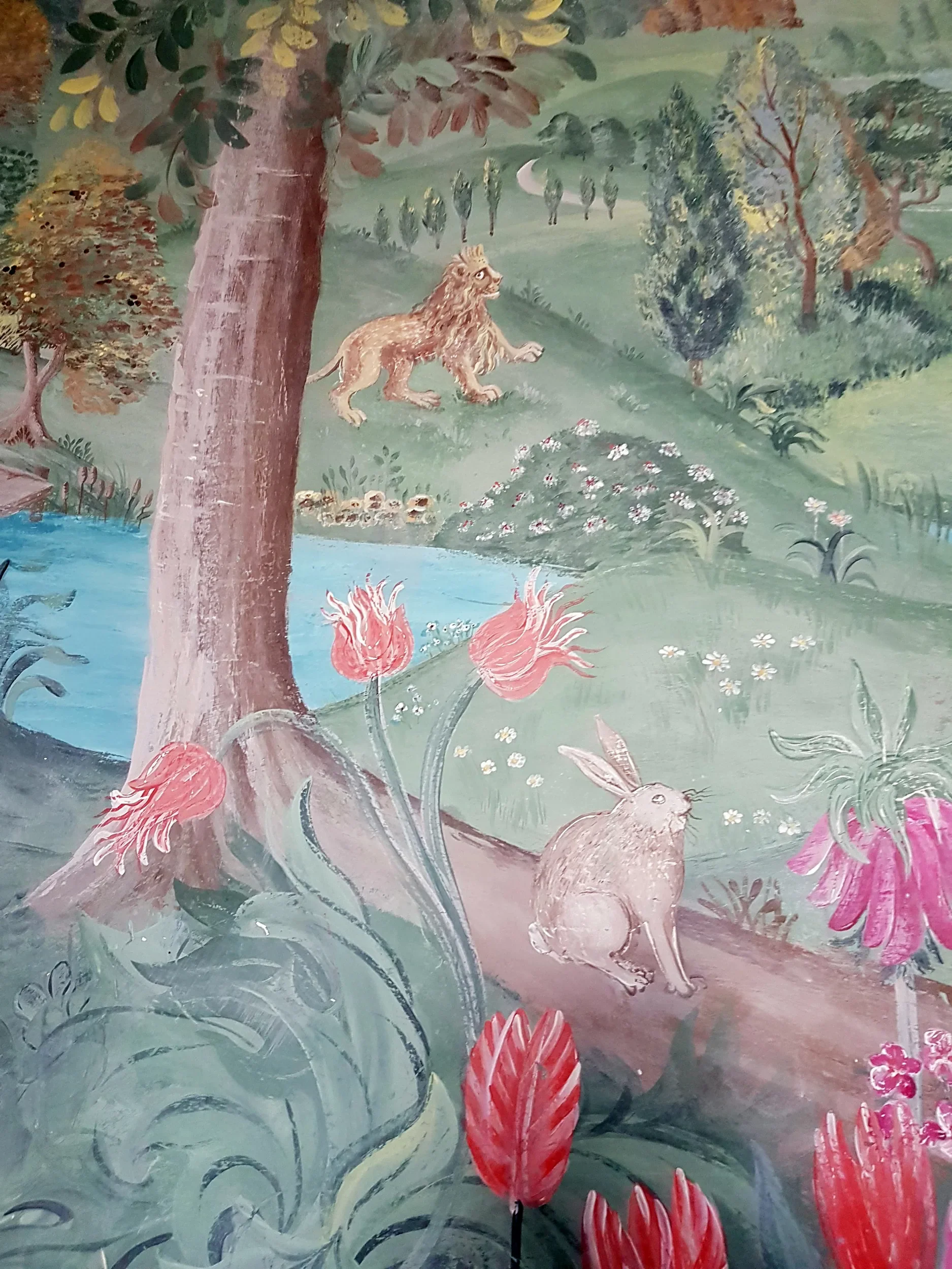 A colorful hand-painted mural depicting a lush natural landscape with a lion sitting on grass, trees, flowers, and a rabbit in the foreground. There is also a pond with a small bird, and rolling hills in the background.