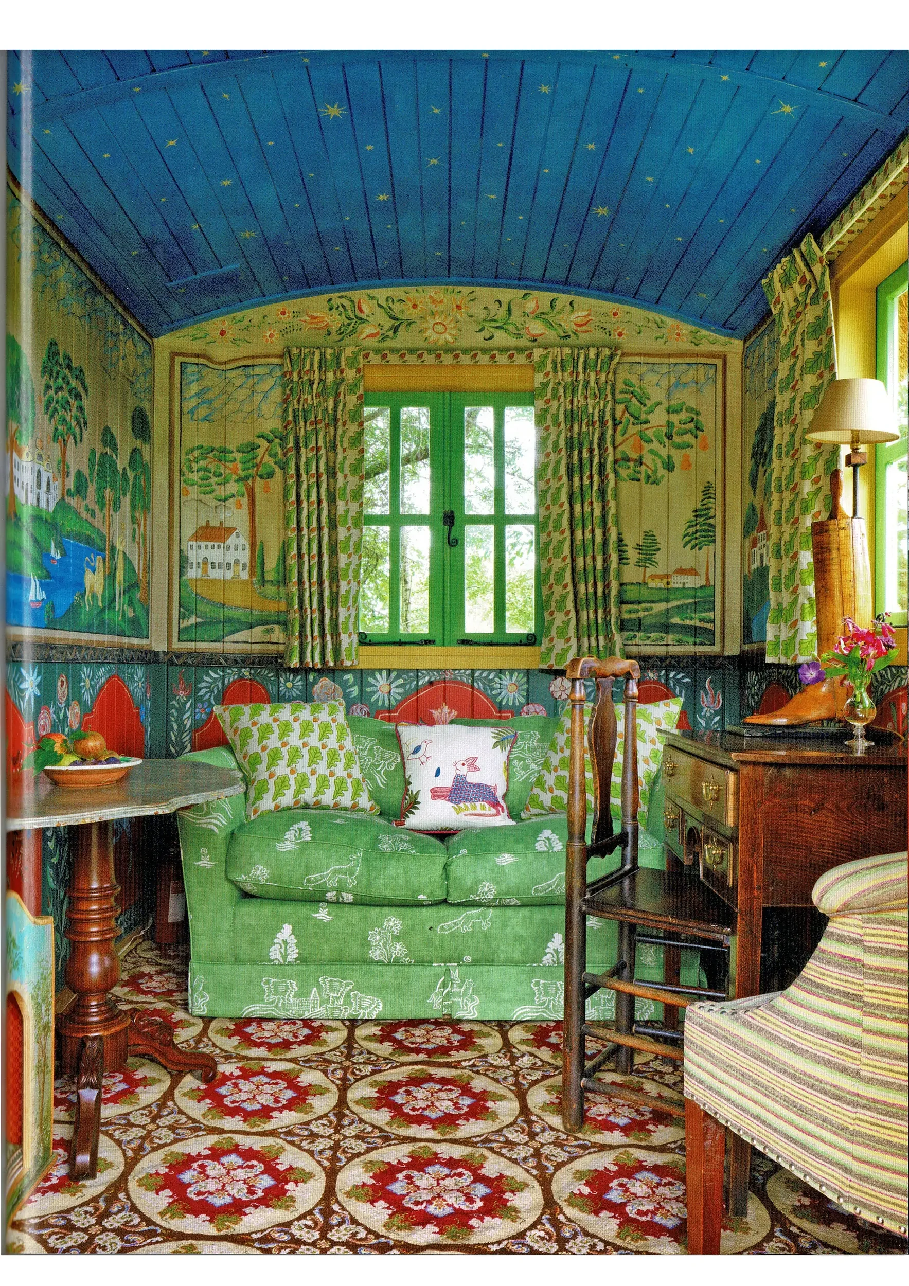 A cozy living room with a vibrant, handcrafted interior. The ceiling is painted dark blue with small yellow stars. The walls feature scenic landscape paintings with trees, houses, and rivers. The window has green trim and floral-patterned curtains. A small green sofa with decorative pillows sits on a red, floral-patterned carpet. Beside the sofa, a wooden side table holds a bowl of fruit. To the right, a striped armchair with a multicolored fabric cushion is visible, and a wooden dresser with a vase of pink flowers stands nearby. A floor lamp with a beige shade is next to the window.