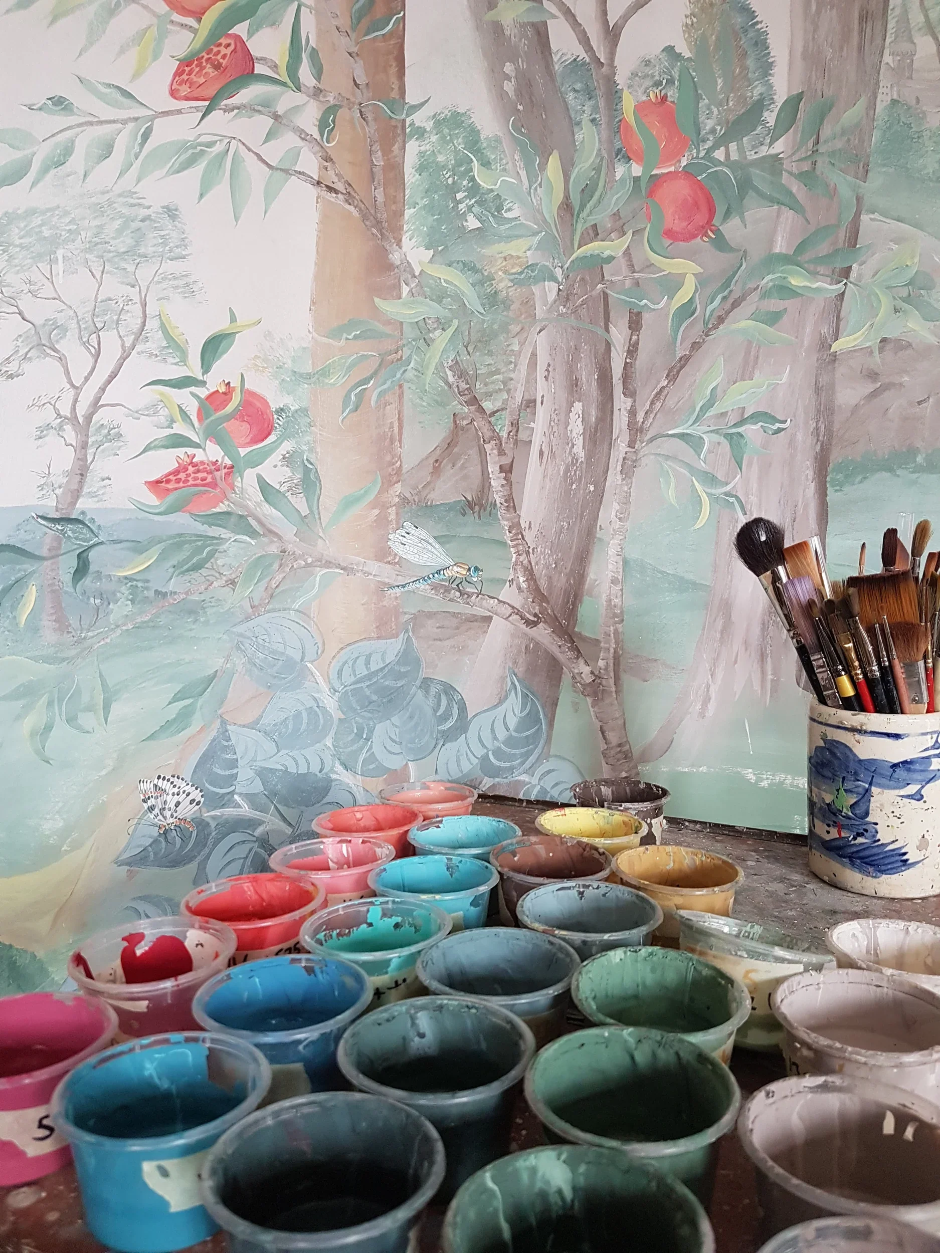 Paint supplies including numerous small containers of various colors of paint, a cup filled with paintbrushes, and a colorful mural in the background depicting trees, pomegranates, and a dragonfly.