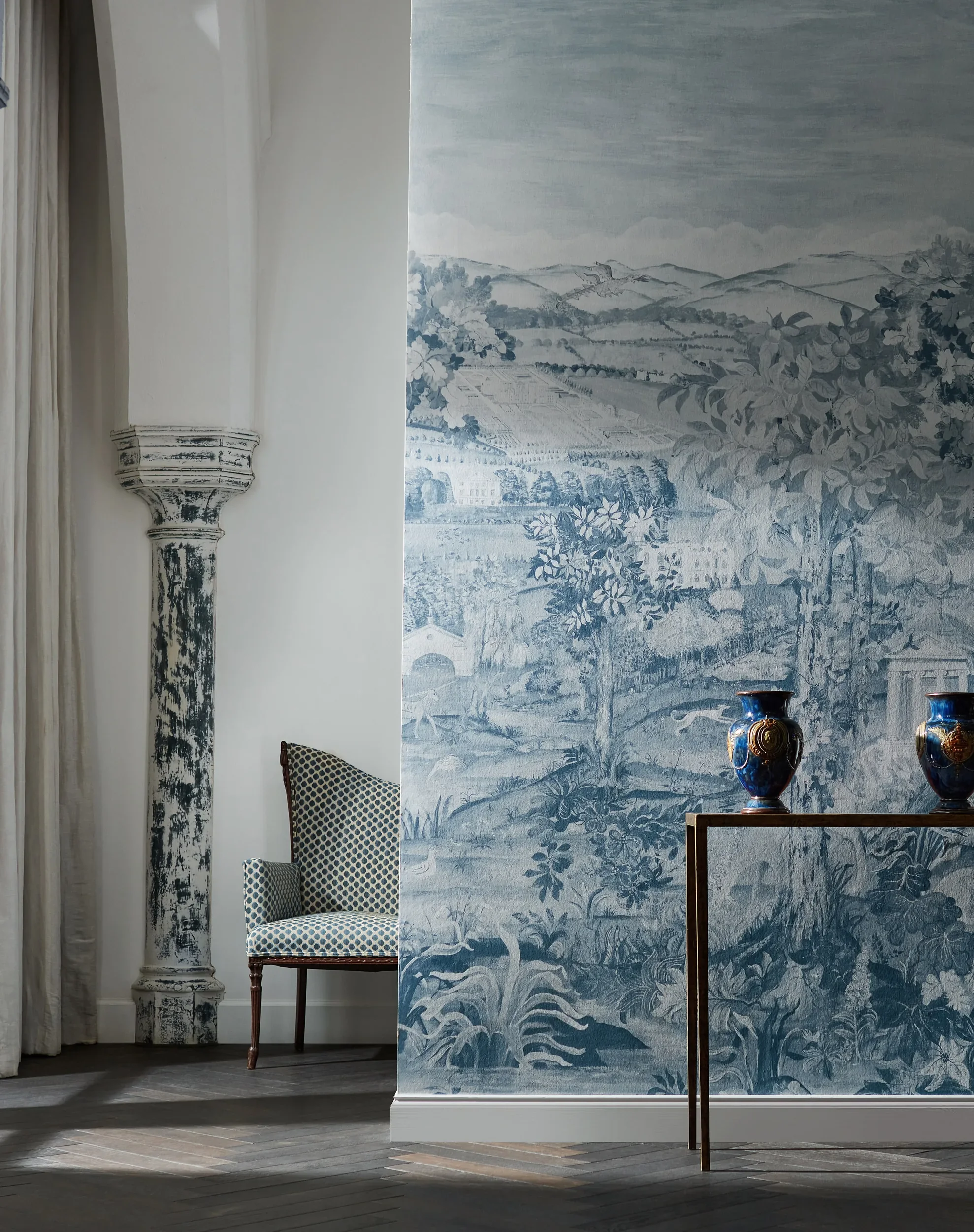 Interior room with a decorative white column, a patterned armchair, a large landscape-style blue and white mural, a wooden table with blue and gold vases, and natural lighting from a window with curtains.