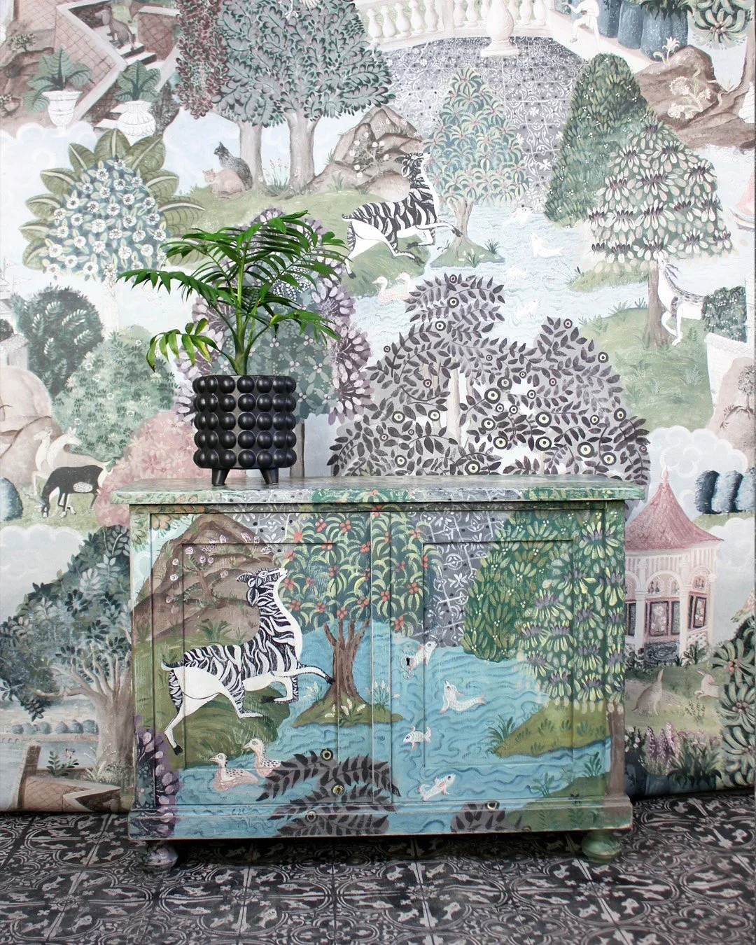 A painted wooden cabinet with a jungle scene of animals and greenery, topped with a black vase holding a green plant, set against a wallpaper with a similar jungle theme.