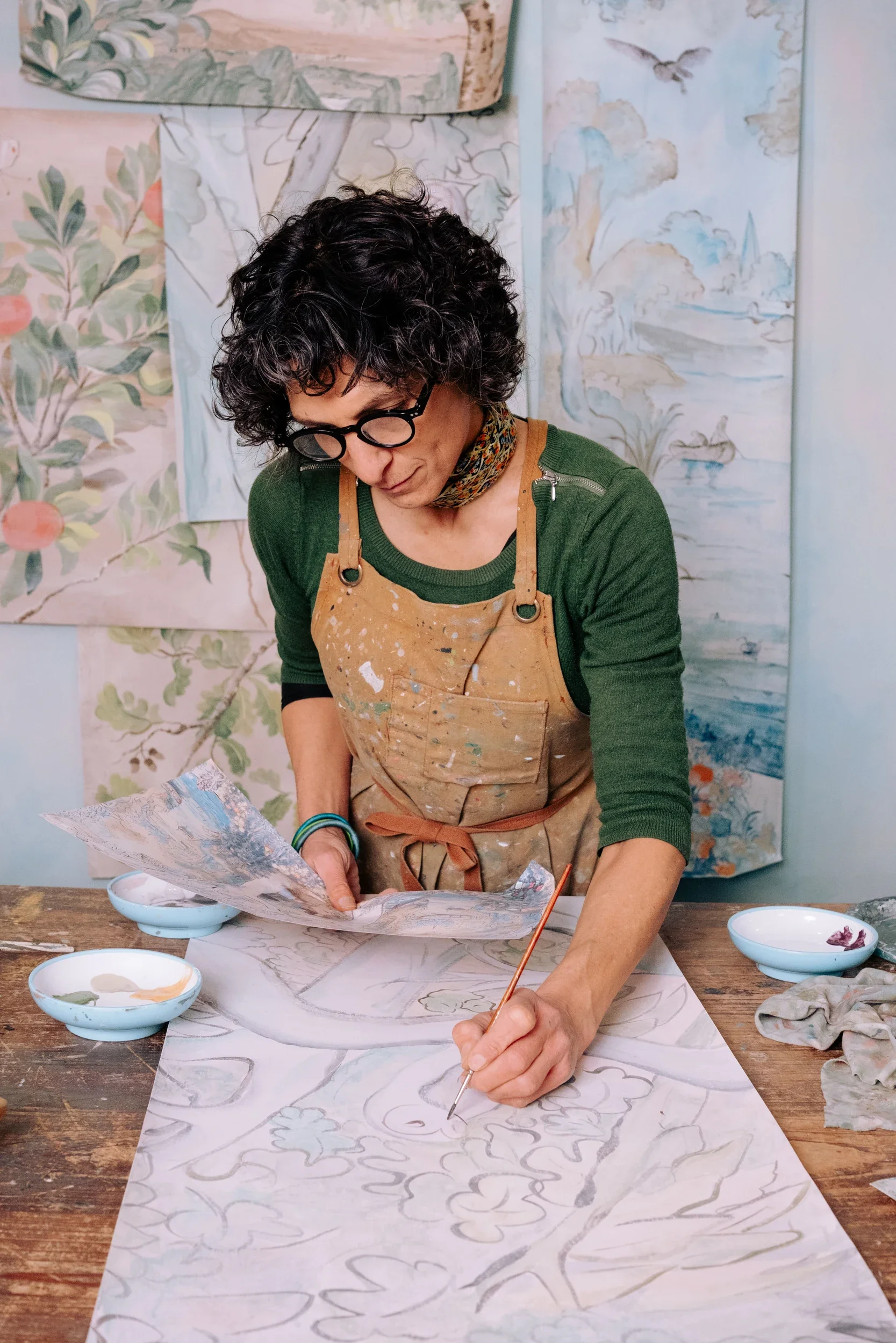 Woman with curly hair, glasses, and a green shirt, working on a large, detailed watercolor painting of a landscape with trees and water, in an art studio with finished landscape art on the wall.