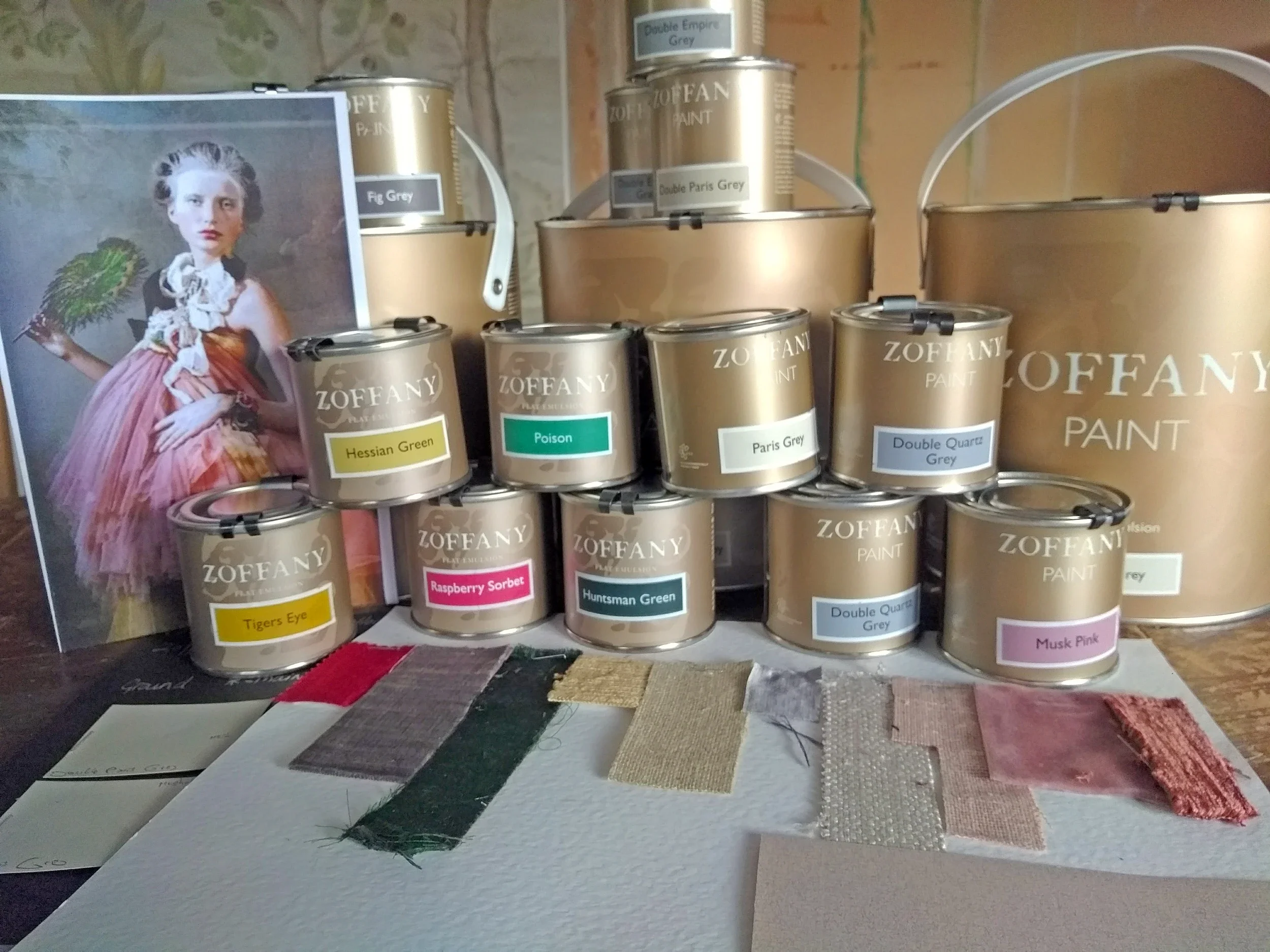 Display of paint cans labeled with various colors and names, such as Hessian Green, Poison, Paris Grey, Raspberry Sorbet, Tigers Eye, Huntsman Green, Double Quartz Grey, and Musk Pink, along with swatches of similarly colored paint and fabric samples.