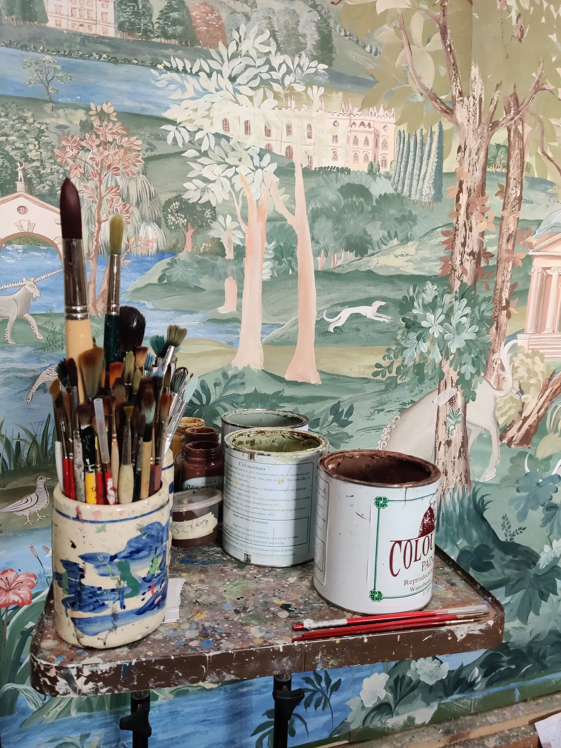 Studio workspace with a table covered in paint cans, brushes, and a colorful landscape mural in the background featuring trees, a river, a castle, and animals.
