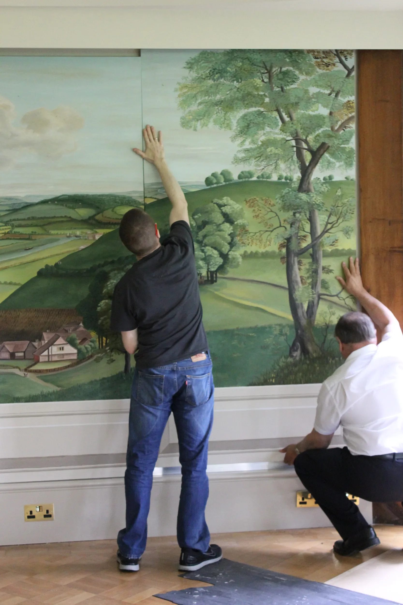 People installing or repairing a large mural or wallpaper with a landscape scene featuring trees, hills, and clouds.