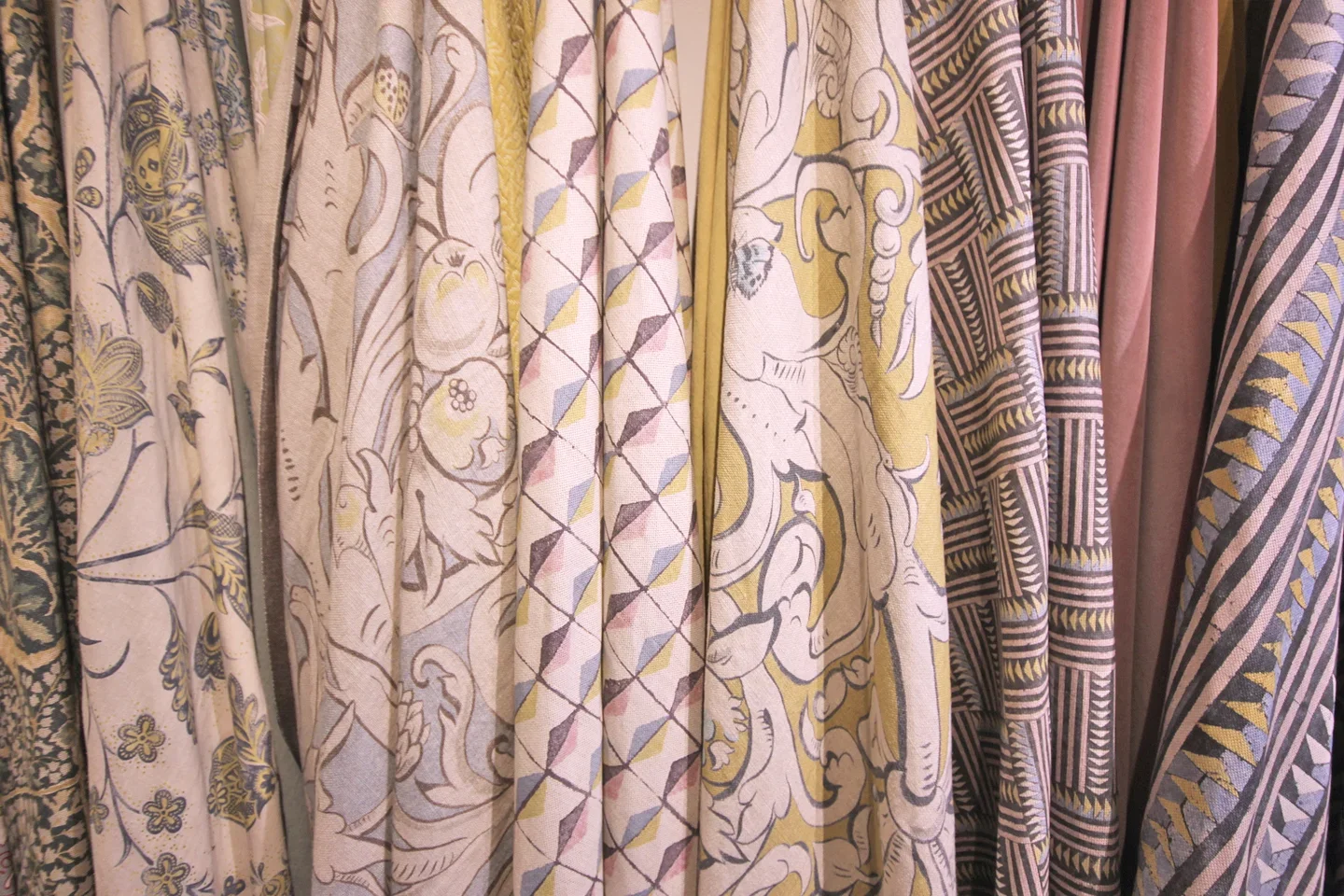 A row of fabric rolls with various patterns and colors on display. Pattern includes Bacchus for Lewis & Wood by Melissa White.