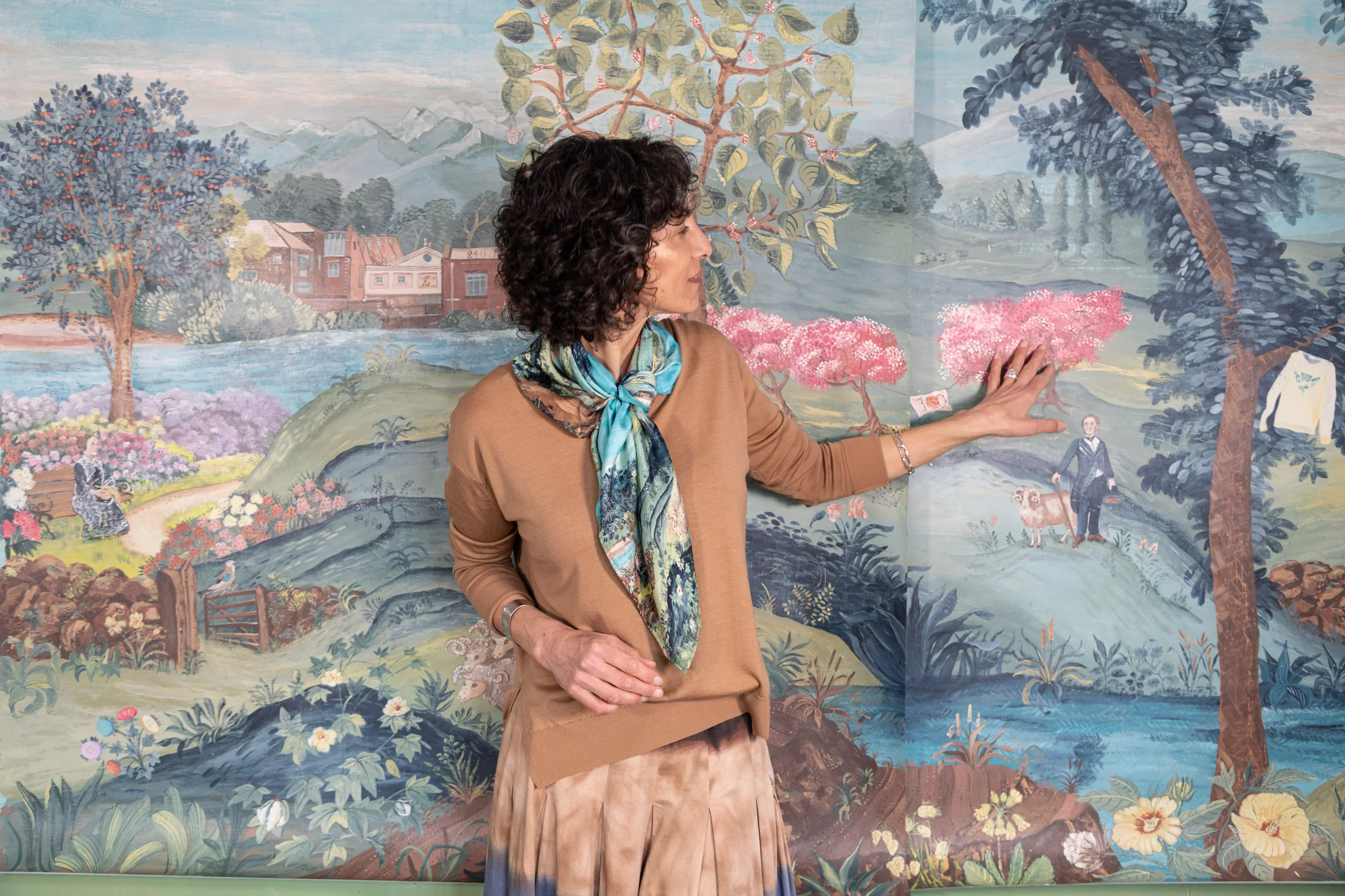 Woman standing in front of a detailed mural depicting a scenic landscape with trees, mountains, a river, and a house, while touching a pink flowering tree.