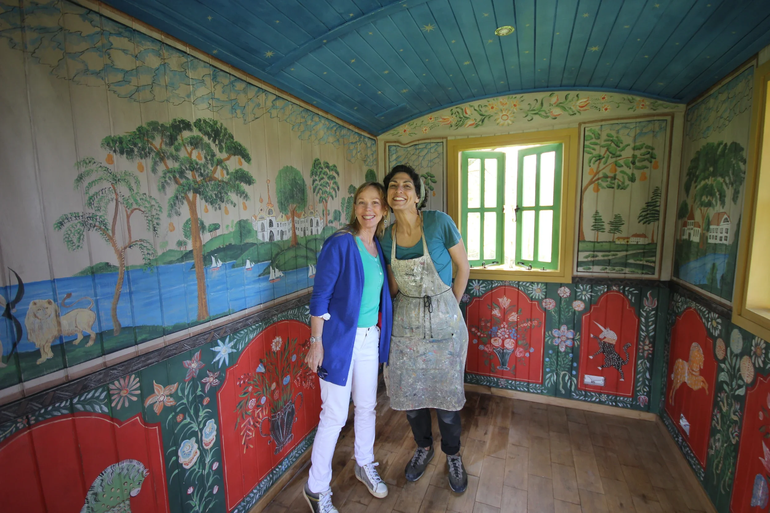 Two women smiling and standing close together inside a colorful, painted room with mural walls and a painted ceiling. One has blonde hair wearing a blue jacket, the other has dark hair wearing an apron and casual clothes. There is a window with open green shutters behind them, revealing bright outdoor light.