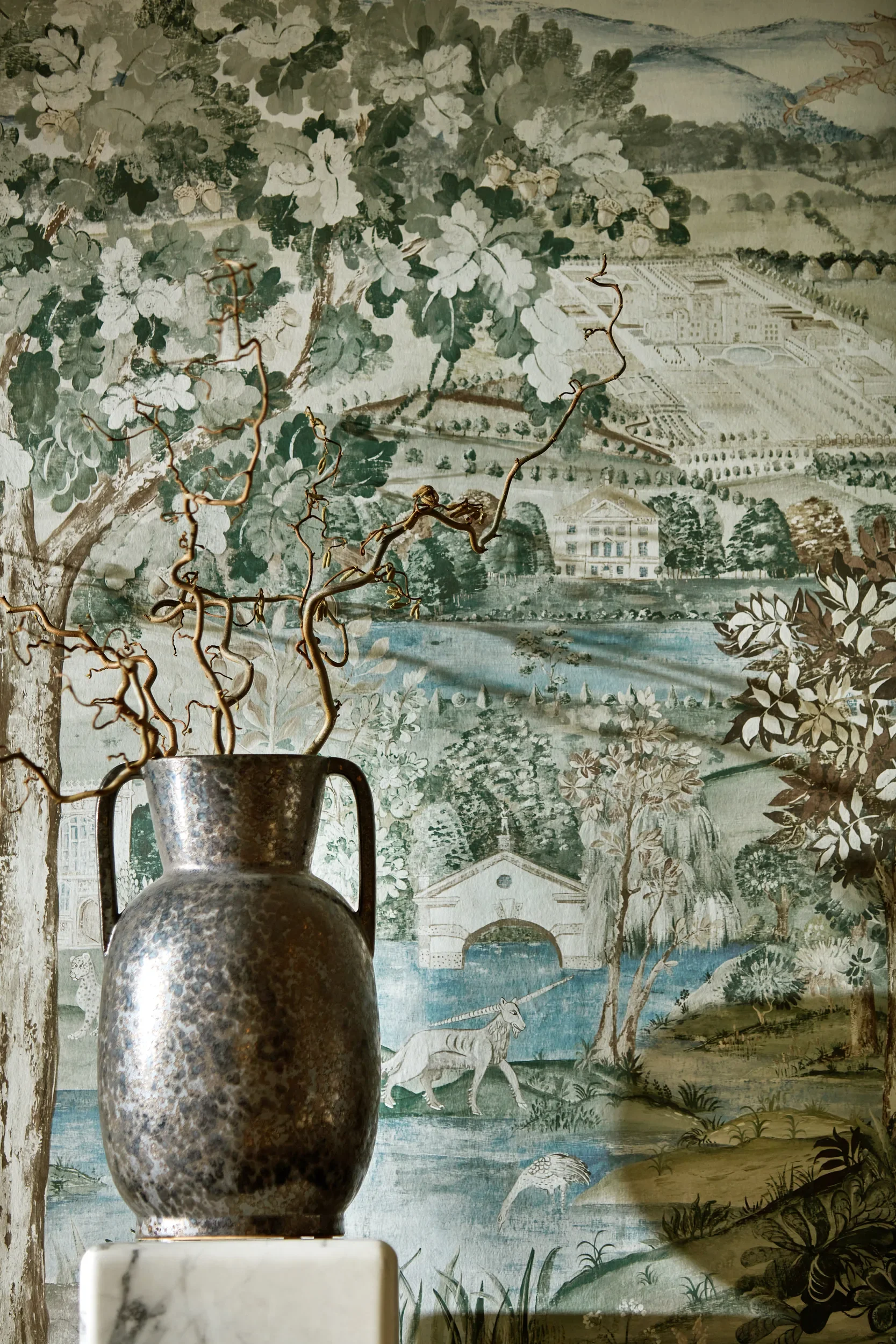 A metal pitcher in front of a detailed mural depicting a landscape with animals, trees, and buildings.
