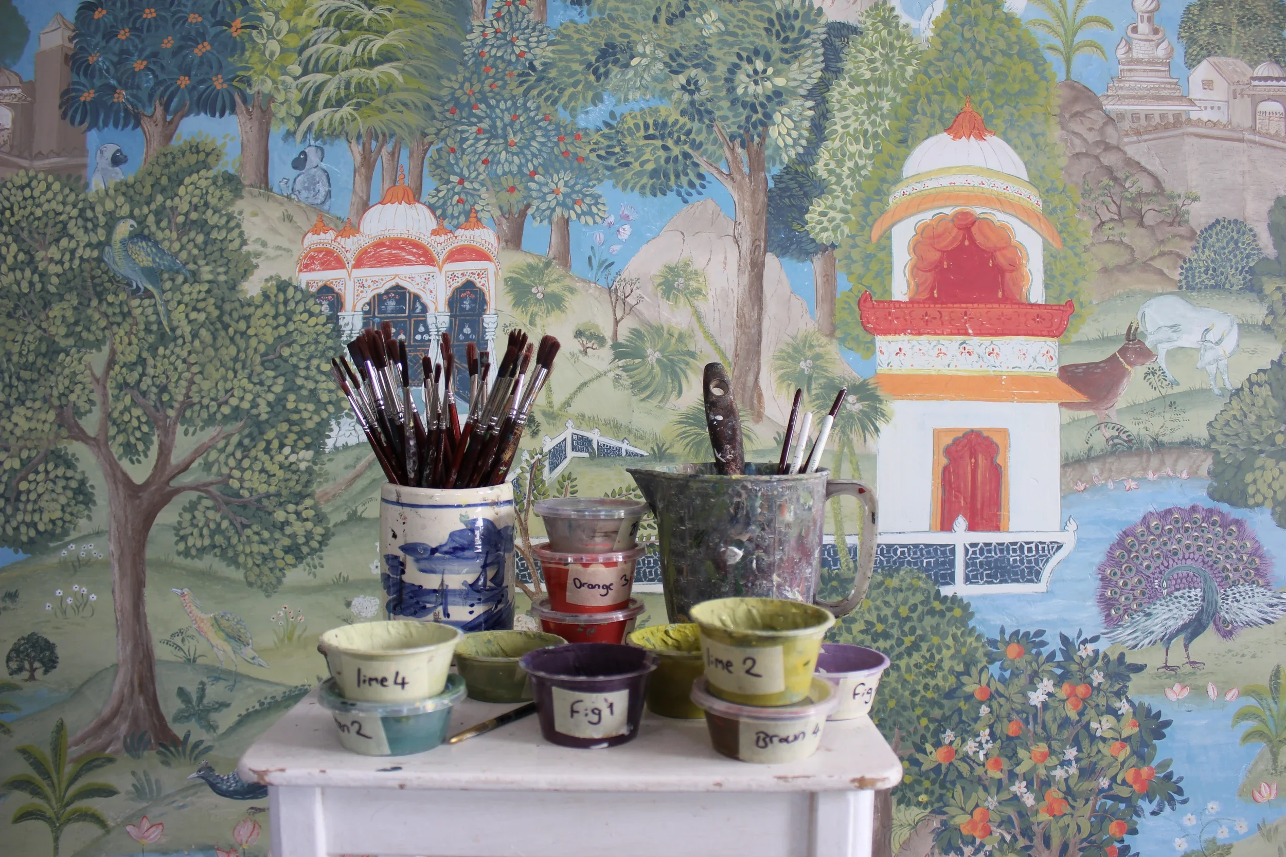 A colorful mural of a lush garden with trees, birds, and buildings in the background. In front, there are multiple artist paintbrushes in containers and several paint cups with labels, indicating different colors or steps in an art project.