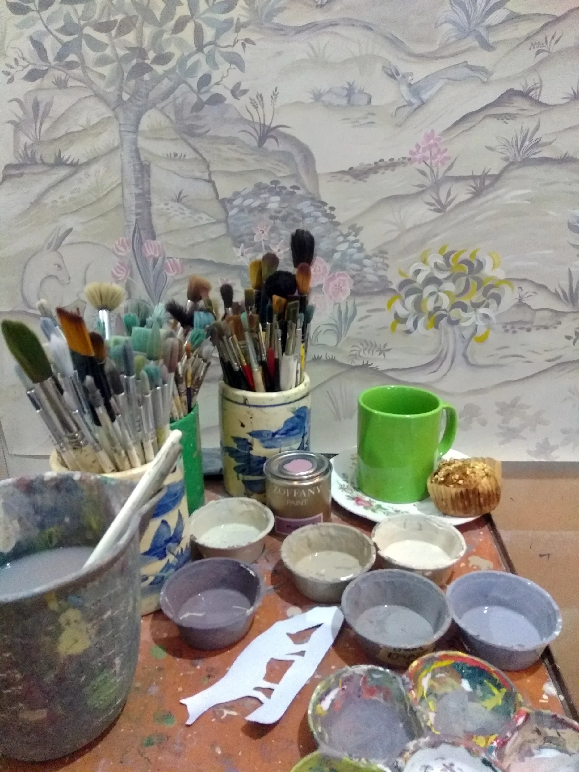 A messy table with paint supplies including brushes, paint cans, and bowls of paint, in front of a large painting of a landscape with animals, trees, and rocks.