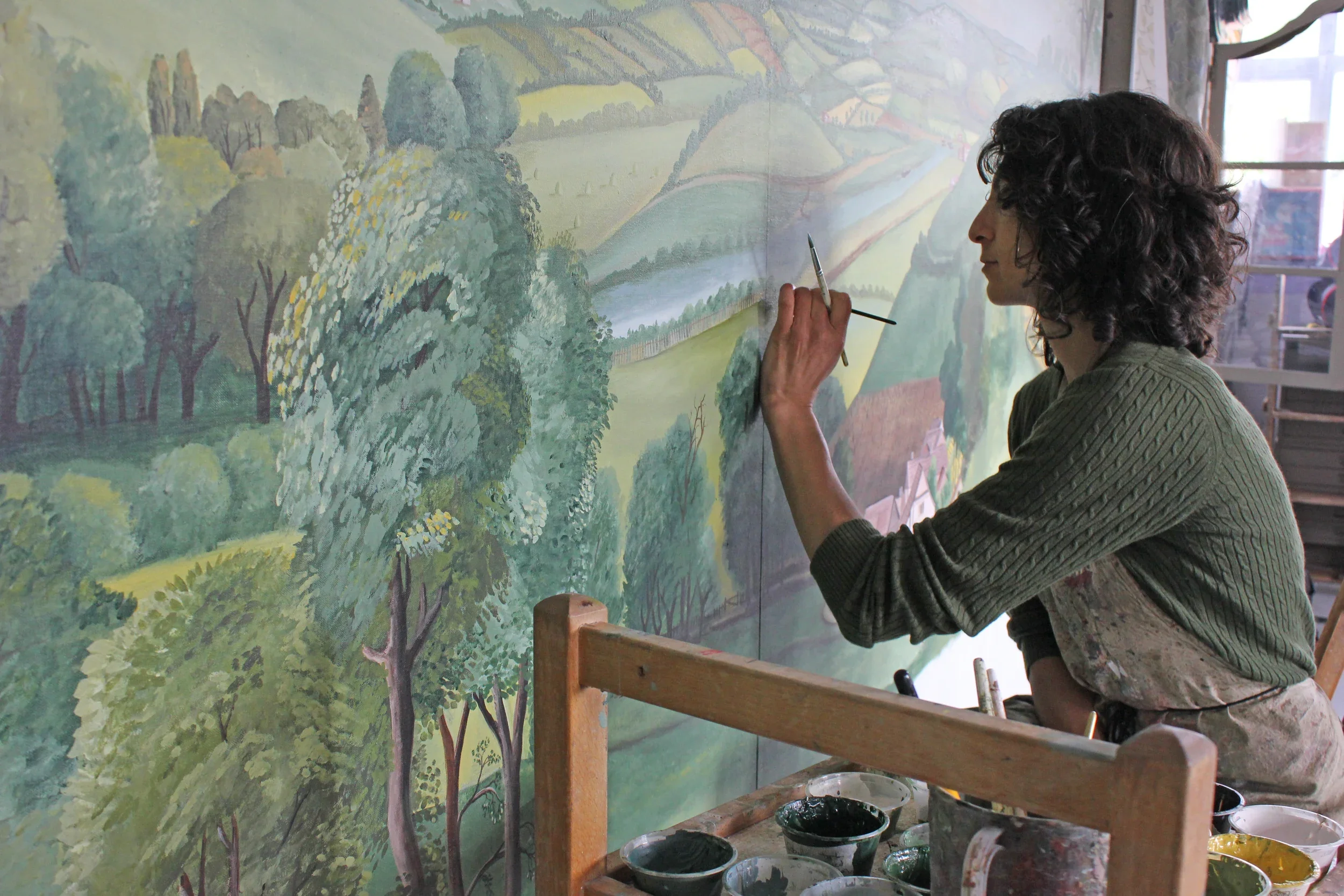 An artist painting a landscape mural in a studio. The mural features a lush green forest, rolling hills, and a small river. The artist is a woman with curly dark hair, wearing a green sweater, standing on a wooden platform with various paint containers nearby.