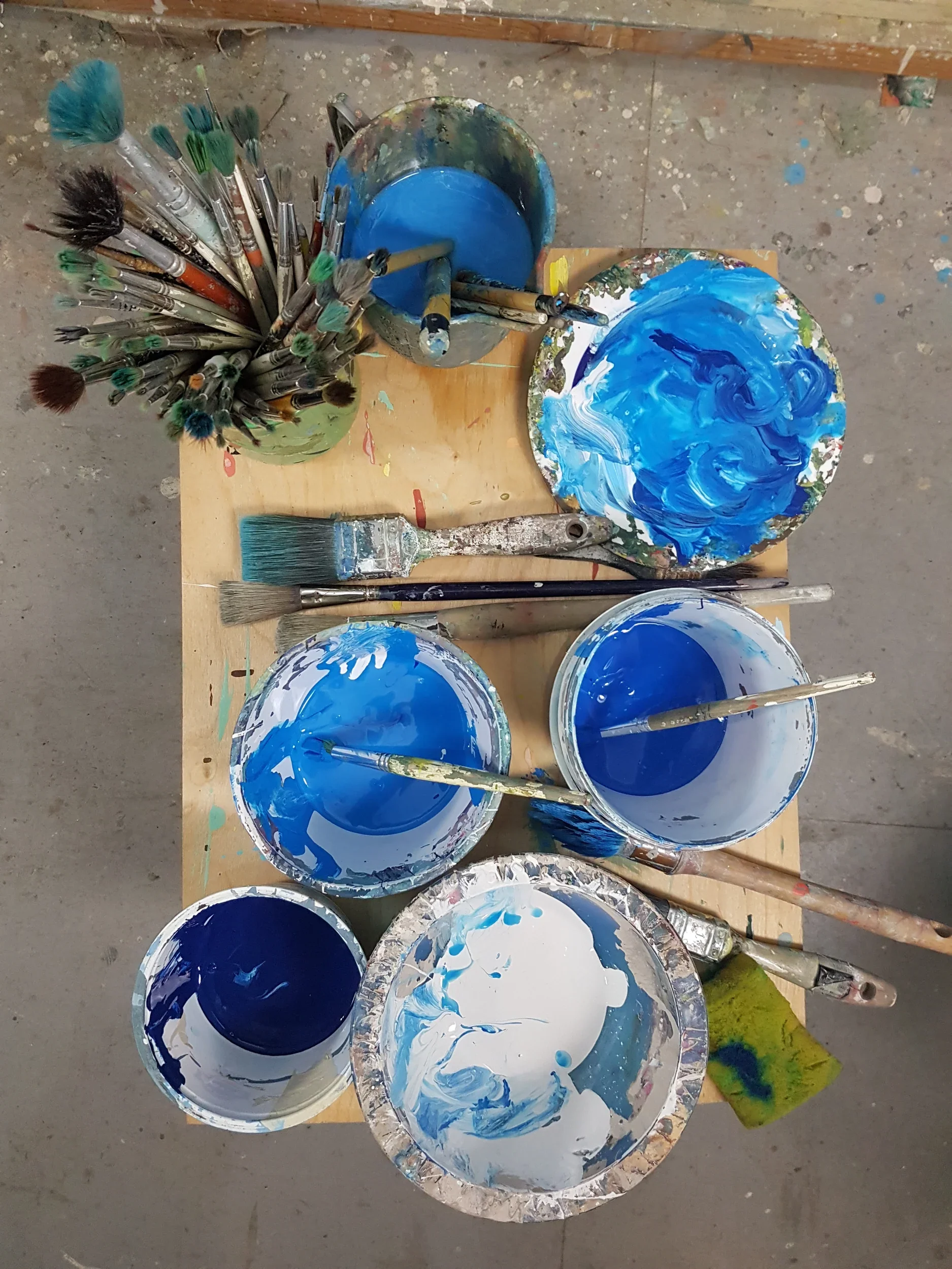 Multiple open cans of blue and white paint with paint brushes resting inside and around them, on a wooden table in an art studio or workshop.