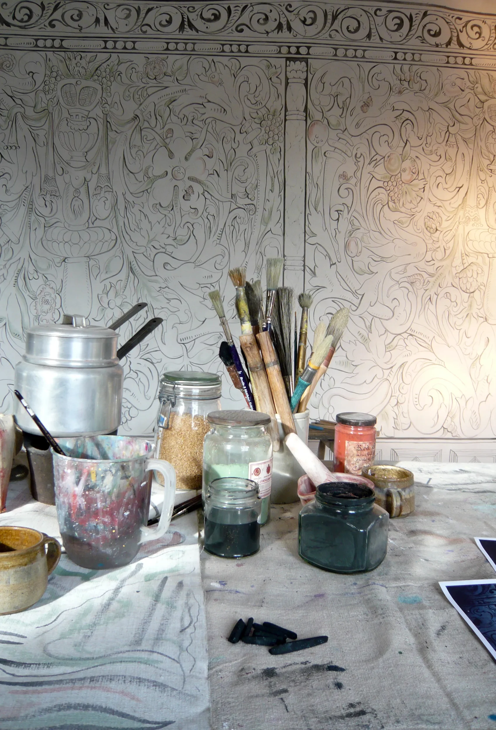An art studio workspace with various jars of paint, brushes, and chalks on a table. In the background, Melissa White's largescale multivine painted cloths for Stiffkey Old Hall, Norfolk.