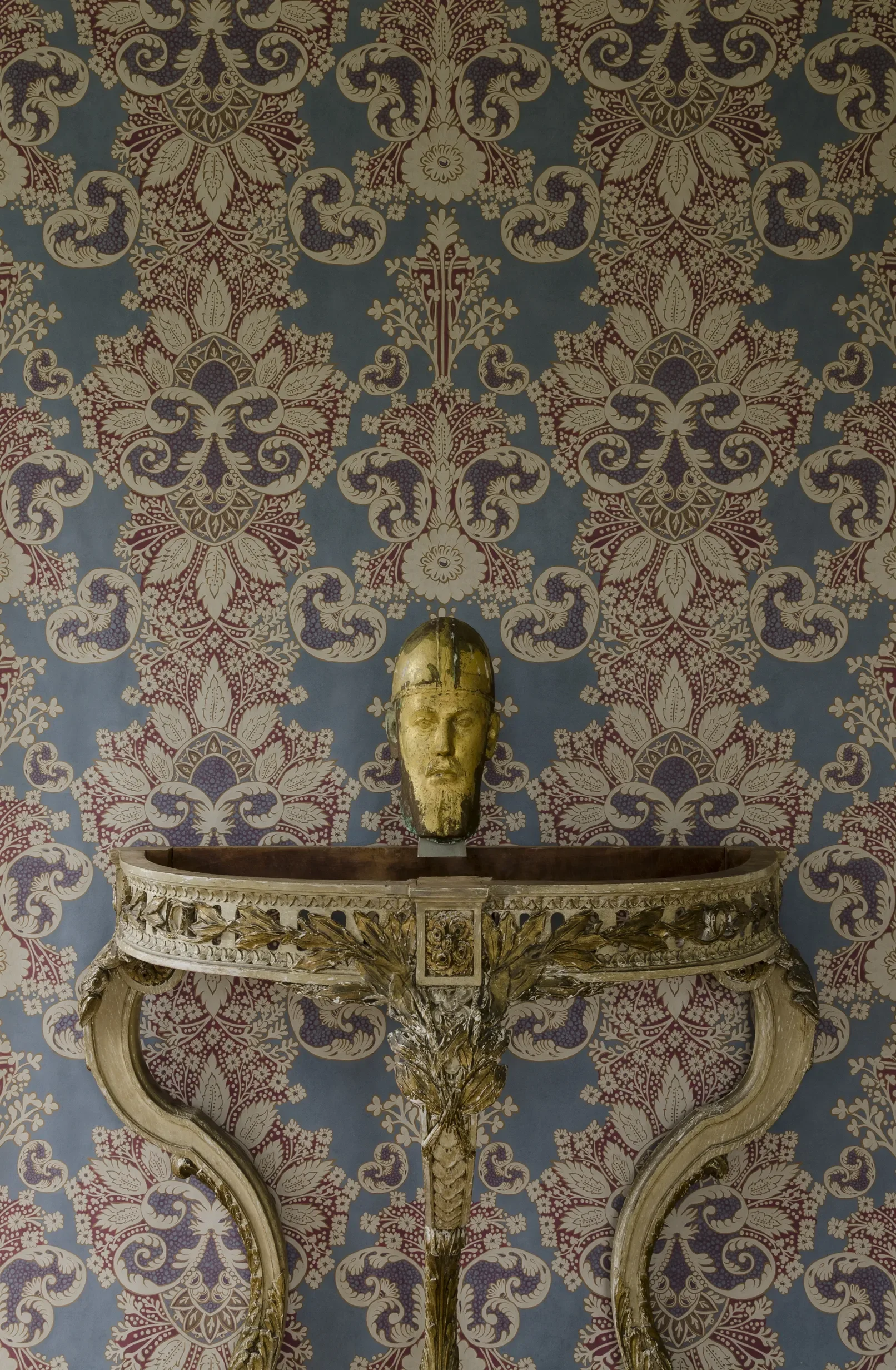 A gold-colored bust of a bearded man on a decorative table against a blue and red ornate floral wallpaper background.