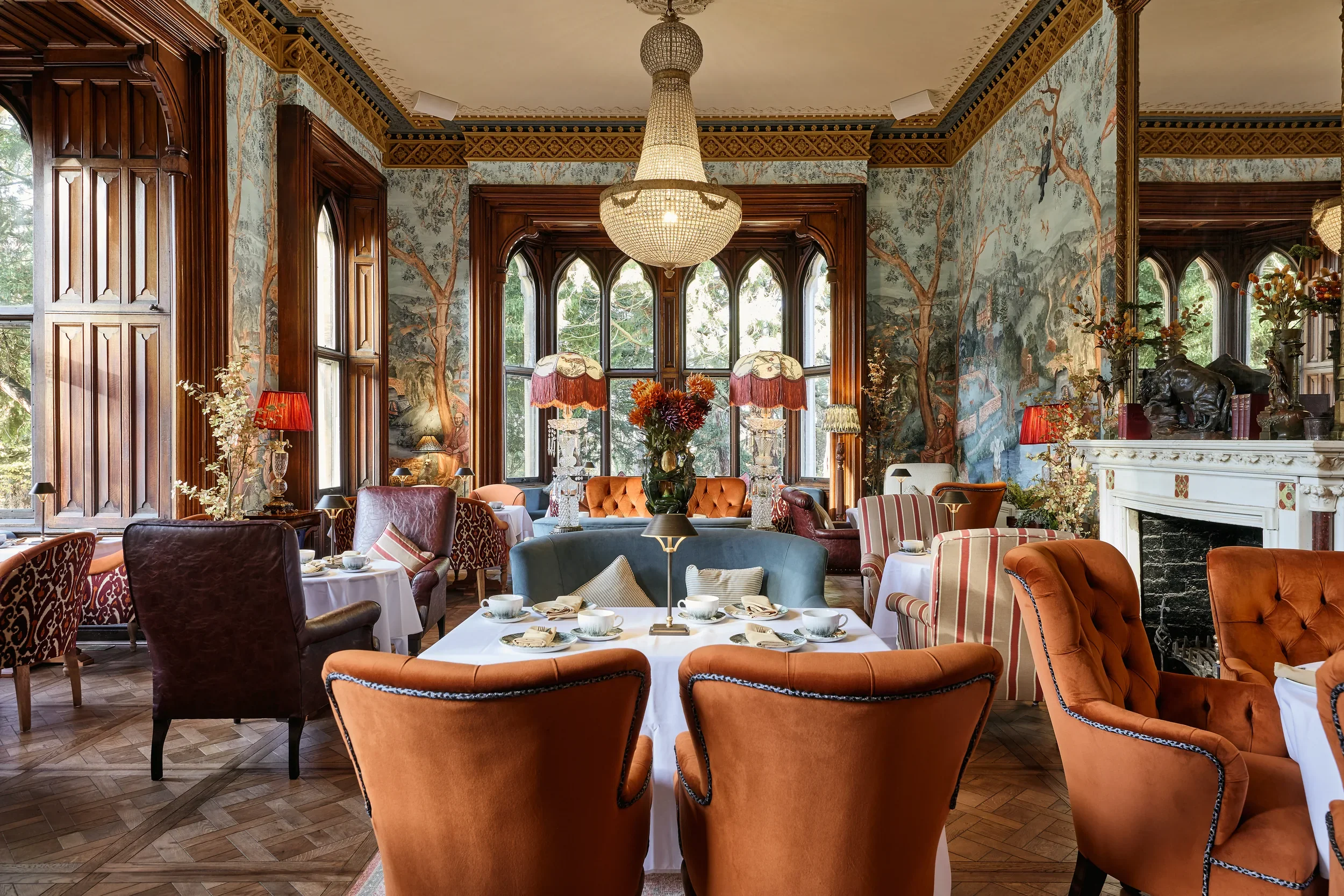 Elegant dining room with large windows, ornate wood paneling, and a mural of trees and landscape on the walls. The ceiling has detailed trim and a large chandelier. The room is decorated with vintage lamps, flower arrangements, and a marble fireplace with sculptures.