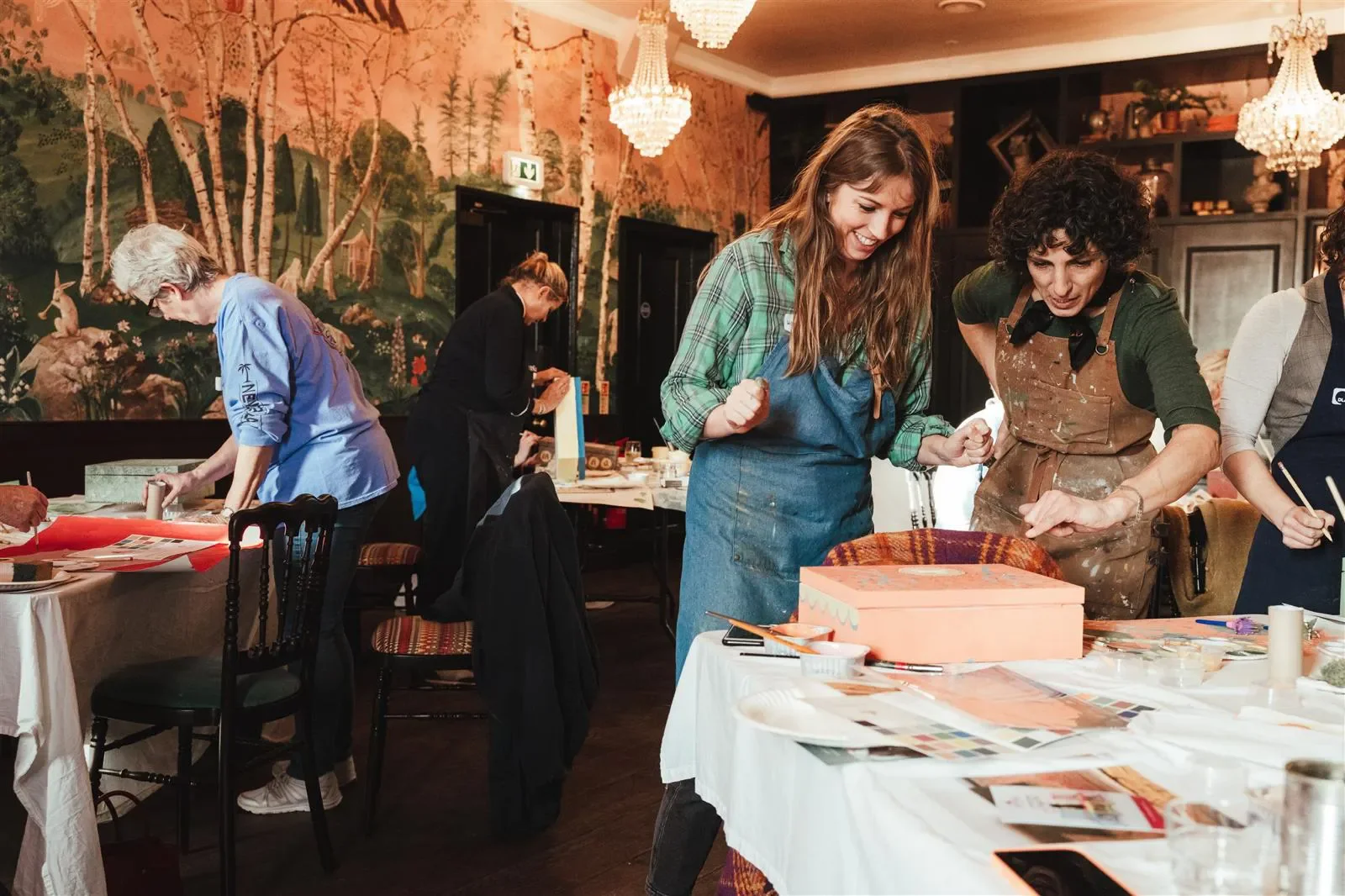 Several women are engaged in an arts and crafts activity at a workshop, with tables covered in papers, paints, and other craft supplies. Two women are in the foreground, smiling and working together, one with long hair wearing a green plaid shirt and apron, and the other with curly hair in a brown apron. The atmosphere is lively and creative, with a decorated wall and chandeliers in the background.