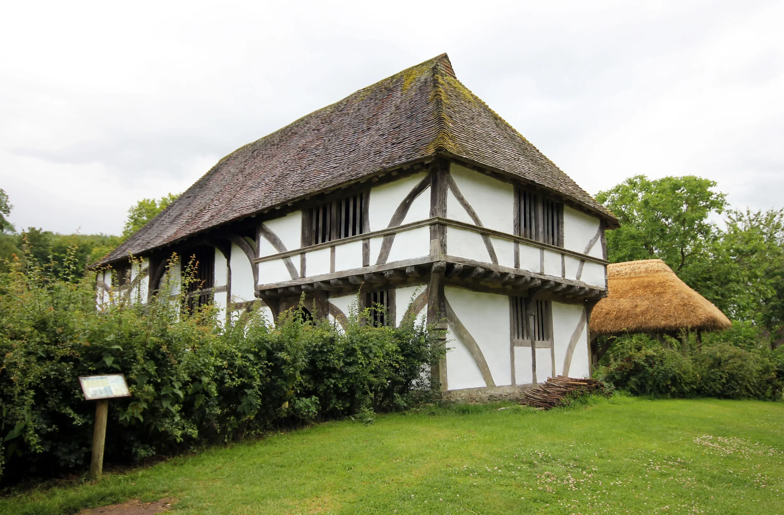 A historic timber-framed building with a steep, moss-covered shingled roof, surrounded by green trees and bushes, with an overcast sky in the background.