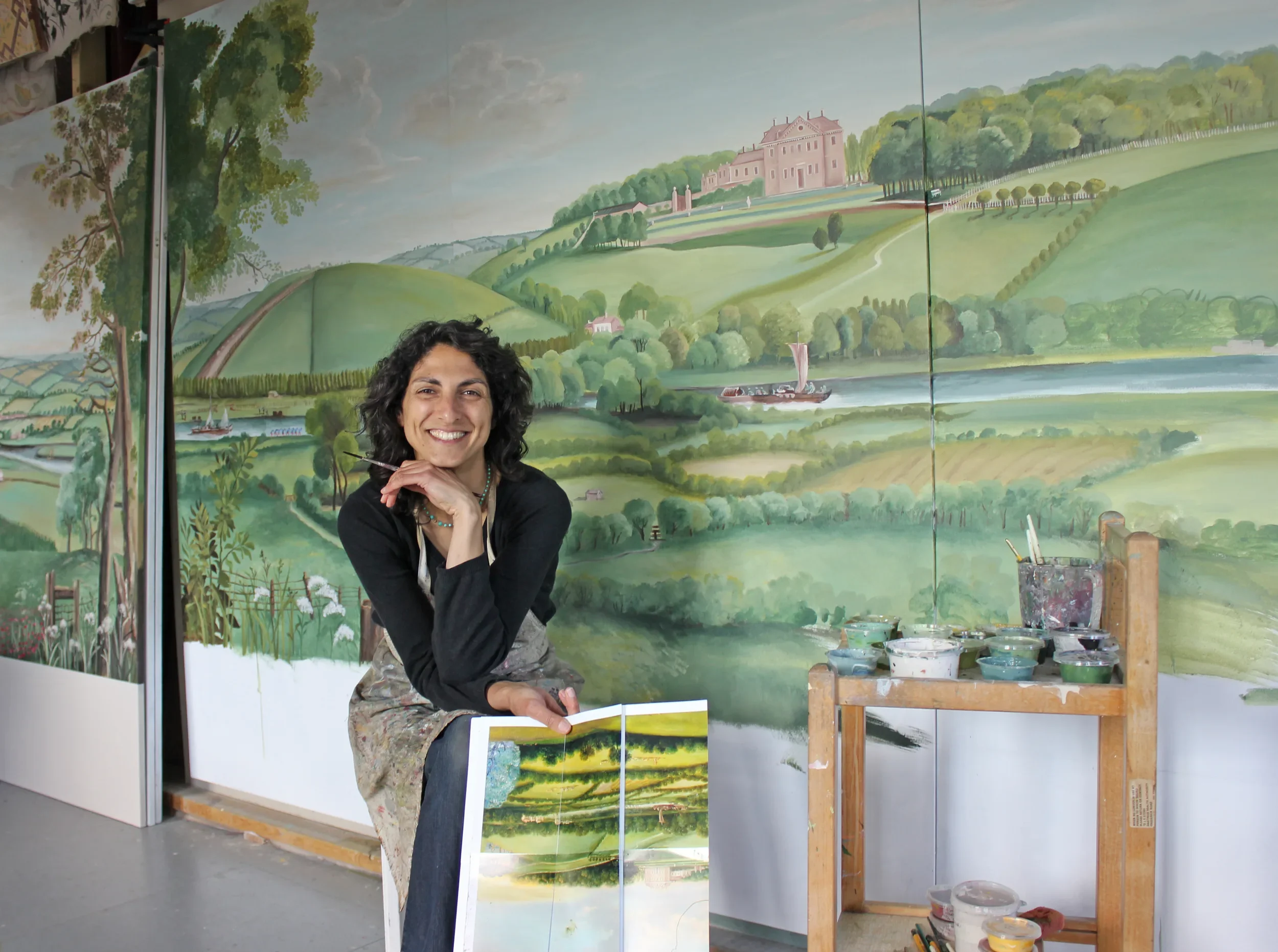 Artist smiling while painting a landscape mural of rolling green hills, trees, a river with boats, and a castle on a hill, in an art studio with painting supplies.