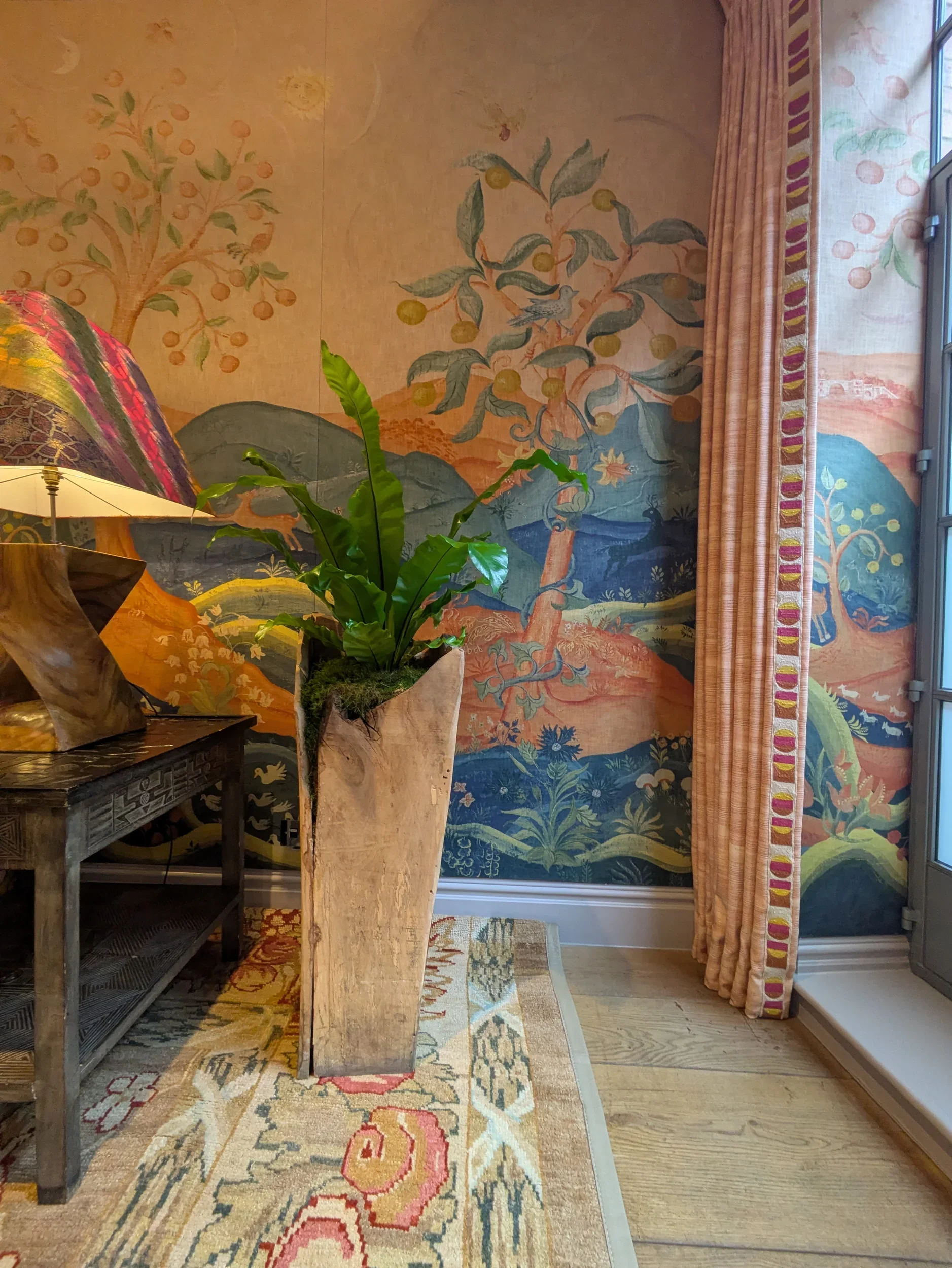 Rufus: Harvest Moon wallpaper by Melissa White for Andrew Martin, room corner detail showing trees, birds, mountains, and sun and moon motifs, with styled interior accessories.