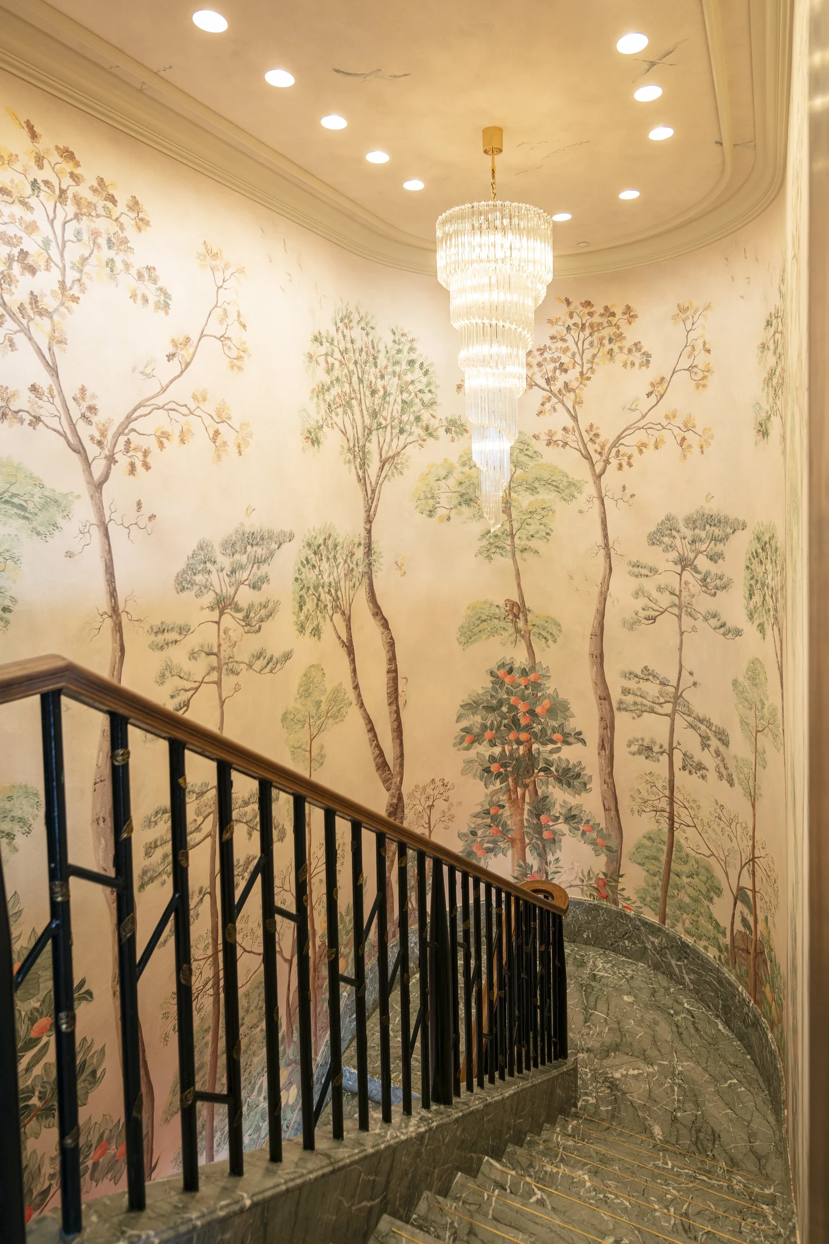 Decorative staircase with a black metal railing and marble steps, against a wall with traditional Asian-style trees and foliage mural. A large chandelier hangs from the ceiling above.