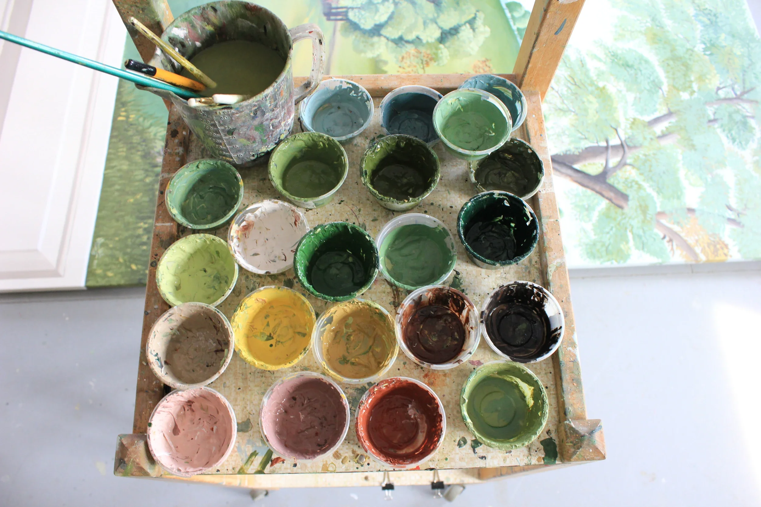 A top-down view of a wooden table with multiple small cups filled with various colors of paint, and a larger container with green paint and brushes inside. There is a painted scenic backdrop visible behind the table.