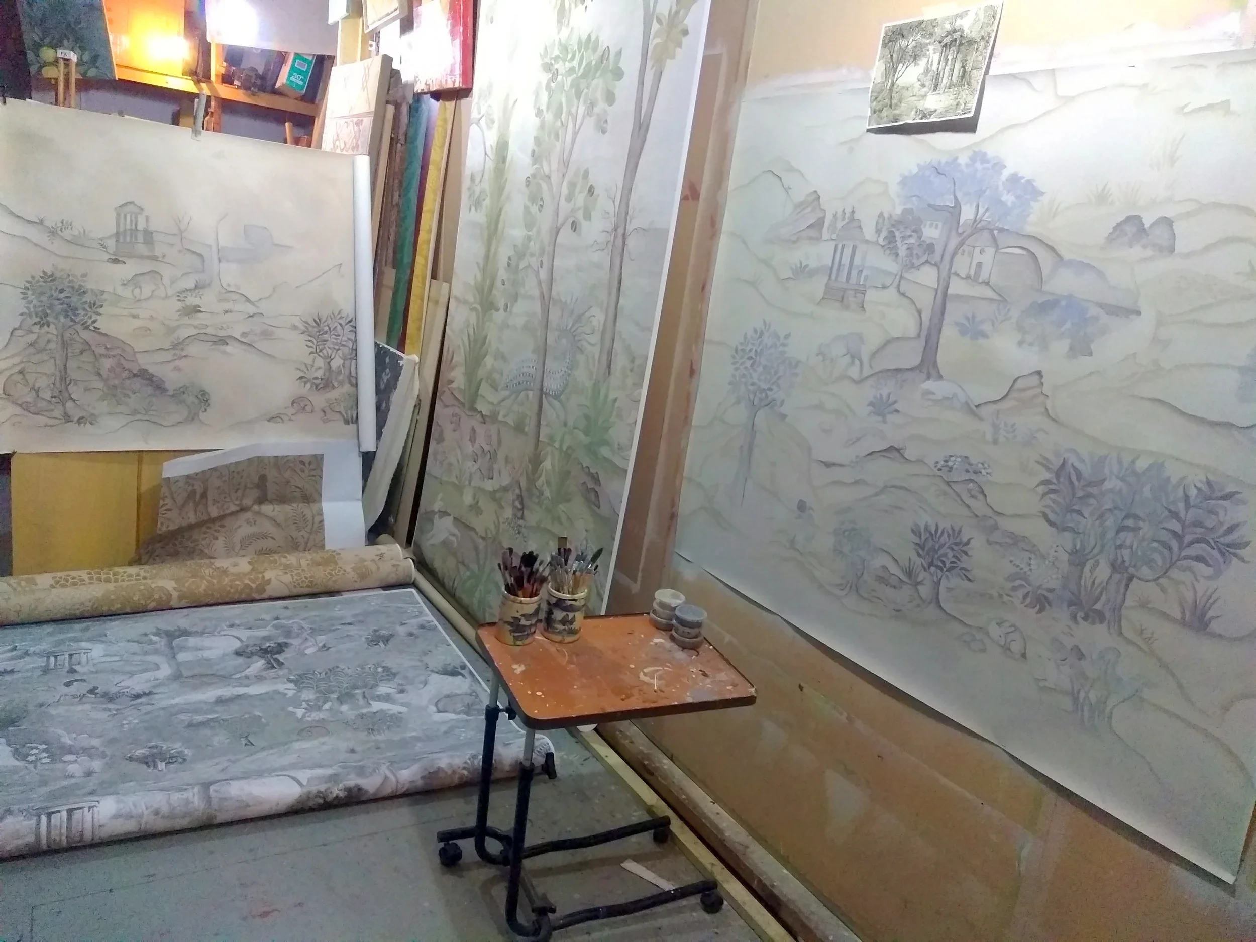 A room with several large landscape paintings leaning against the walls. The paintings depict detailed nature scenes with trees, mountains, and rivers, created with a soft color palette. Art supplies, such as brushes and containers, are on a small table, and a chair is visible with a partially rolled-up canvas on the floor.