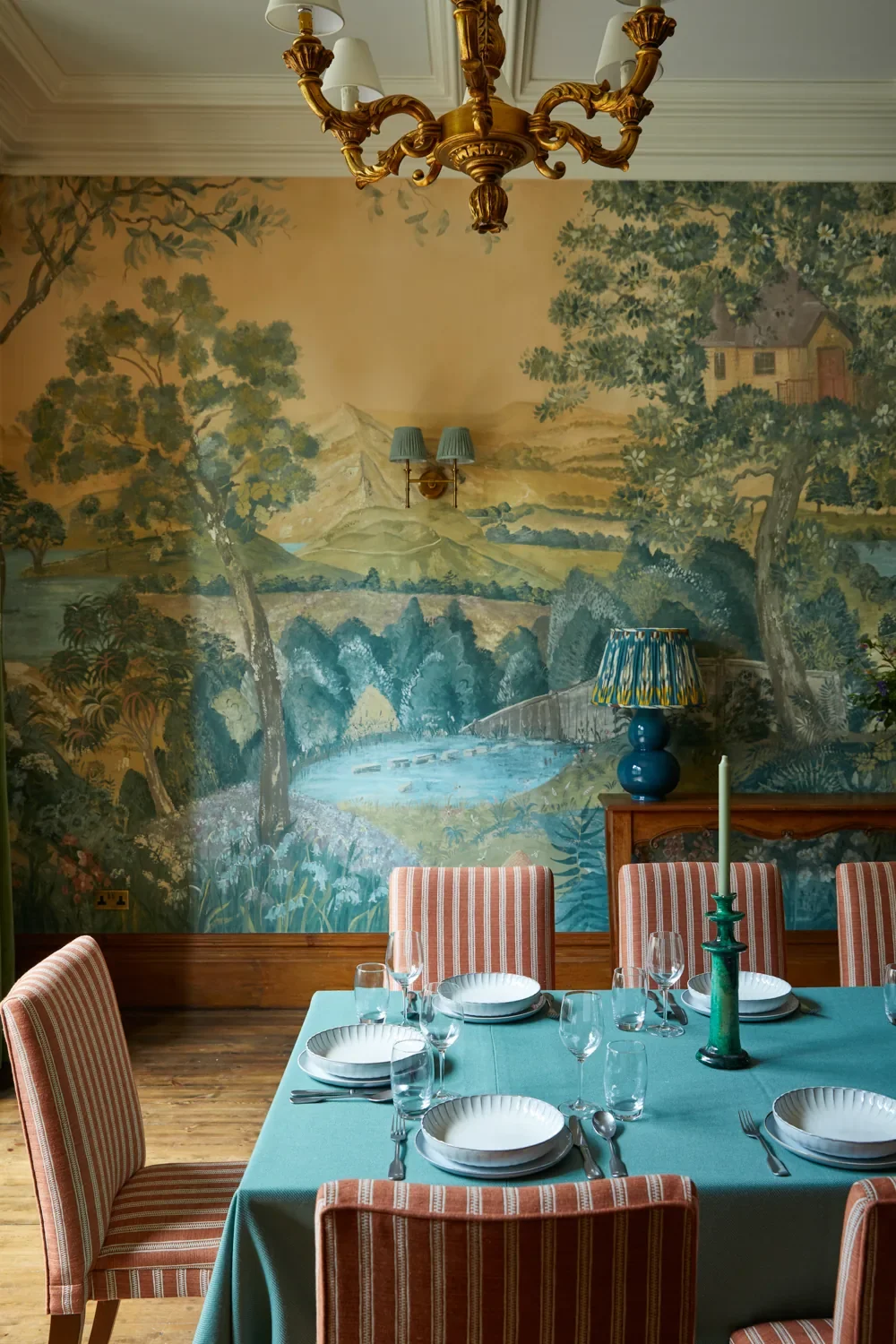 A dining room with a mural depicting a scenic landscape of trees, mountains, and a house, a chandelier hanging from the ceiling, a table set with plates, glasses, and silverware, pink striped chairs, a blue vase-shaped lamp, and a green candlestick with a white candle.