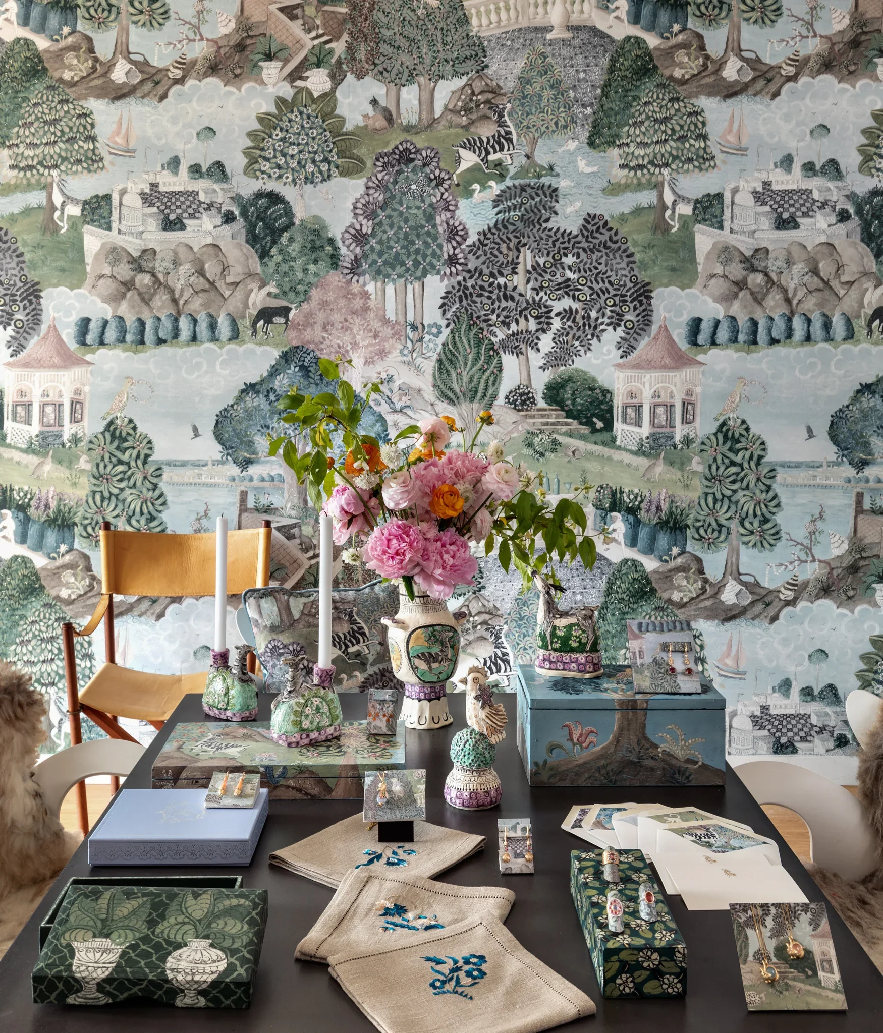 Display of decorative boxes, embroidered cloths, jewelry, and ceramic vases on a table in front of a large wallpaper mural depicting a lush garden scene with trees, animals, and classical architecture.