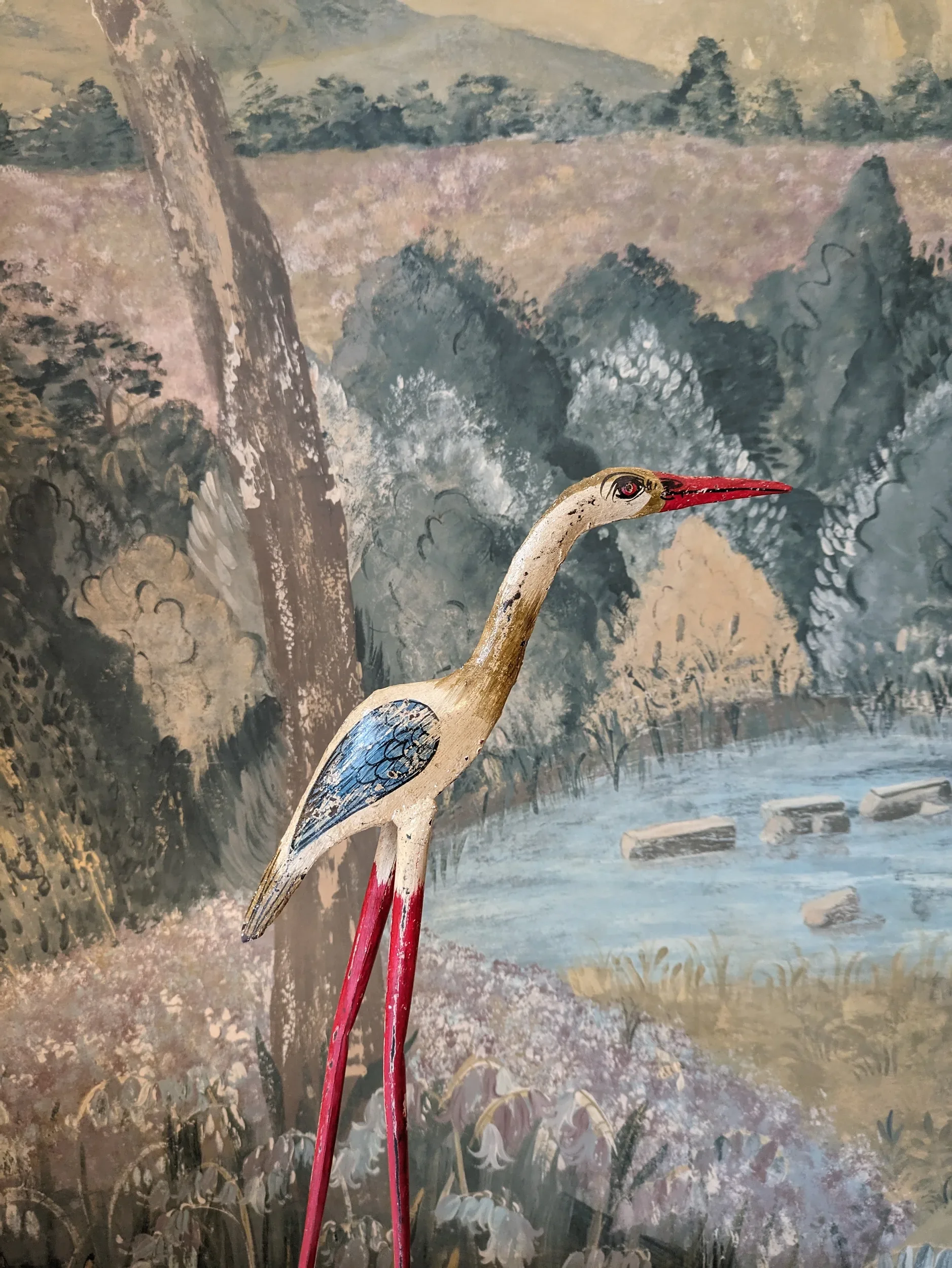 A painting of a landscape with trees, mountains, and a river, featuring a wooden heron sculpture with a long red beak in the foreground.