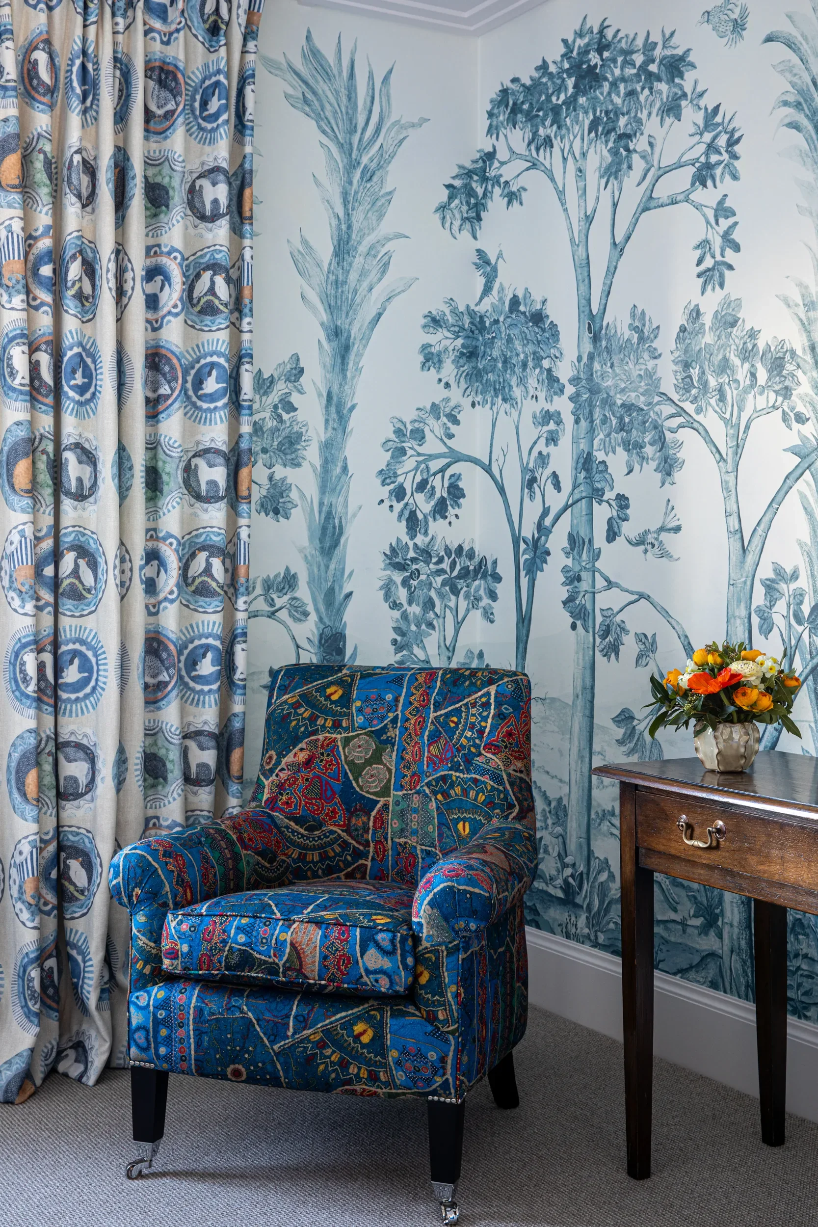 Tall Trees wallpaper by Melissa White for GP&J Baker, Kit Kemp Wallpaper collection, in the delft blue colourway. Shown is an upholstered armchair, a wooden side table with a vase of orange and yellow flowers, and blue and white patterned curtains.