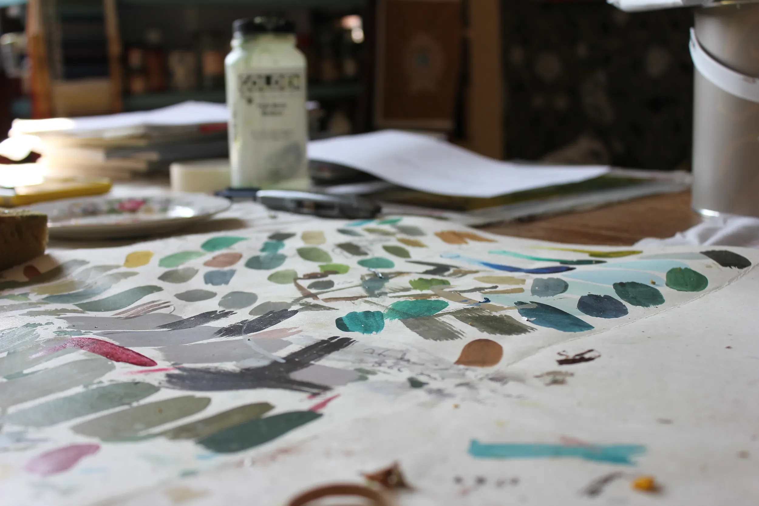 Close-up view of an artist's workspace with a paint palette, papers, and a bottle of paint or medium on a table, with a blurry background of a studio or art room.