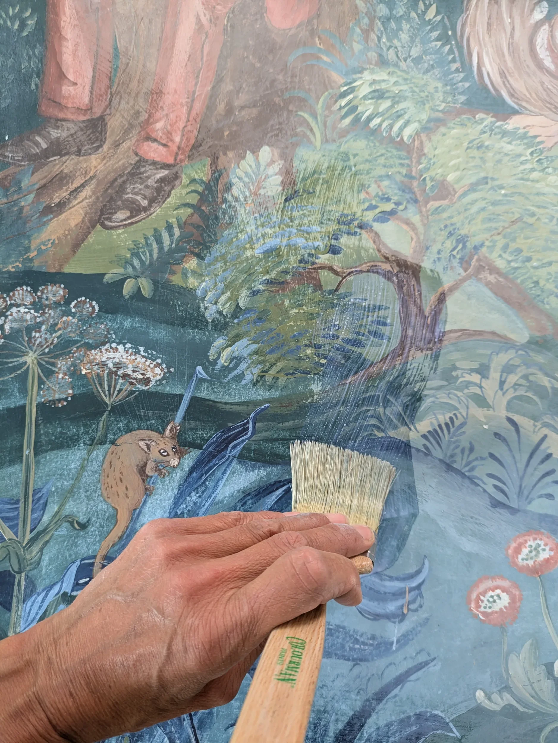 A person holds a brush while painting a colorful, detailed scene of a lush landscape with trees, flowers, and a small animal.