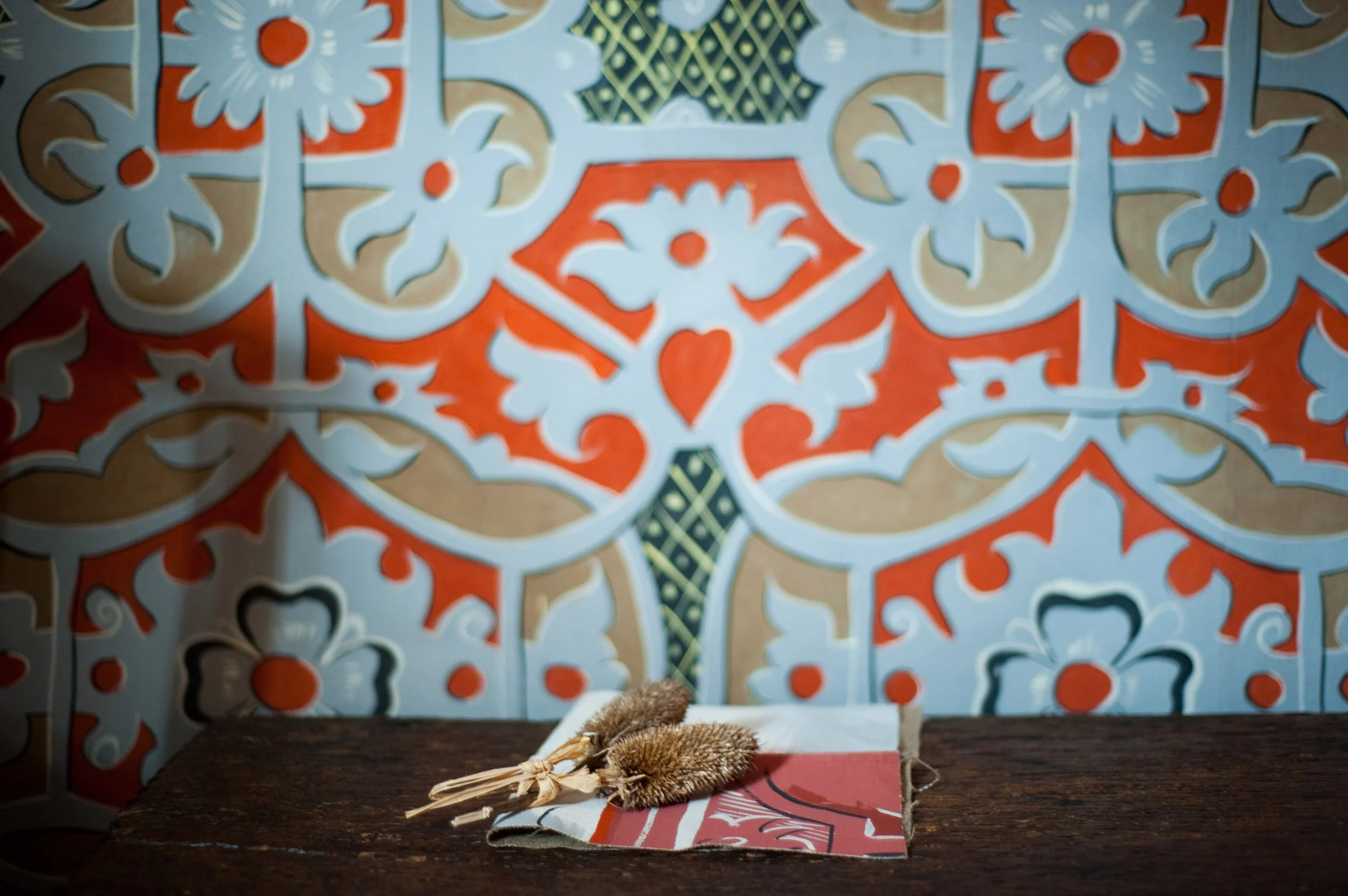 Dried plants on a book on a dark wooden table in front of a decorative tile wall with intricate patterns in blue, orange, beige, and black.