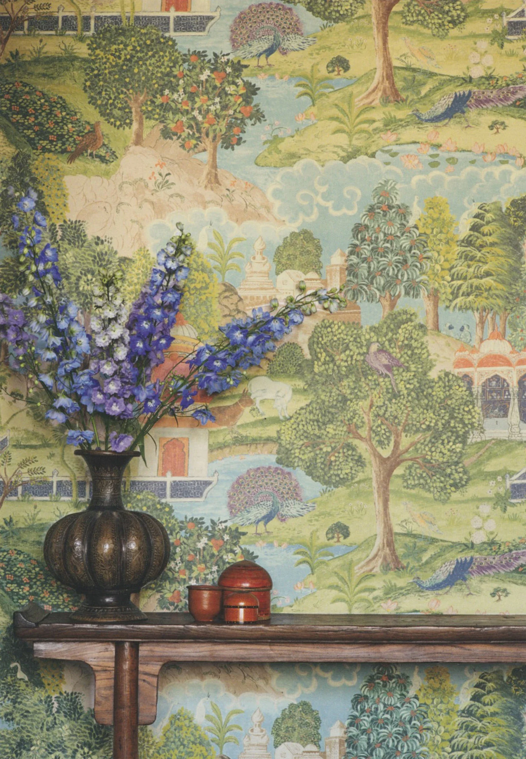 A decorative background featuring a traditional Asian style painting with peacocks, trees, and water, with a dark wooden table in front holding a black vase with purple flowers and three small wooden containers.