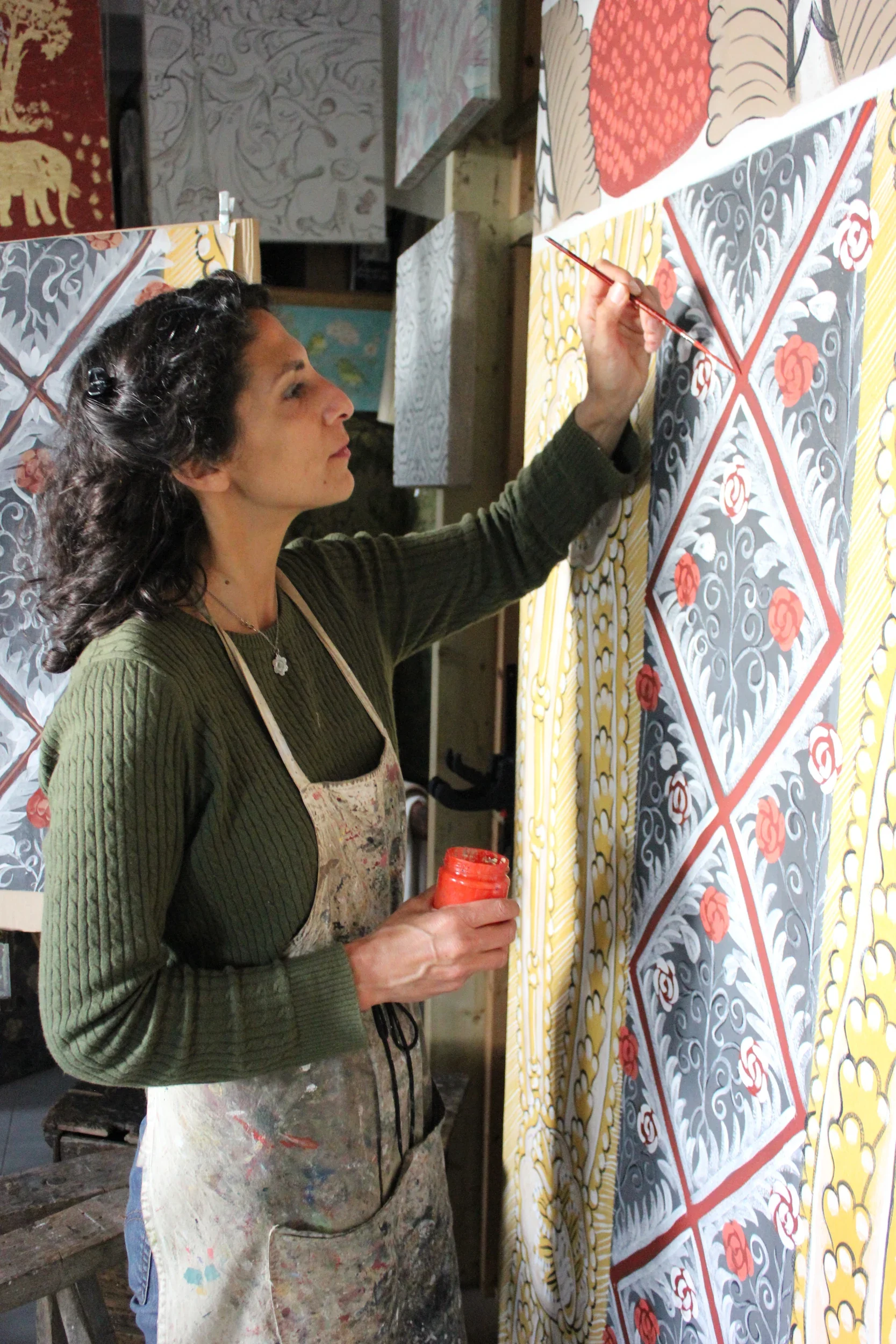 A woman painting intricate floral and geometric patterns on a large canvas with a small paintbrush in her right hand, holding a jar of orange paint in her left hand. She is wearing a green sweater and a paint-splattered apron, with dark, curly hair.