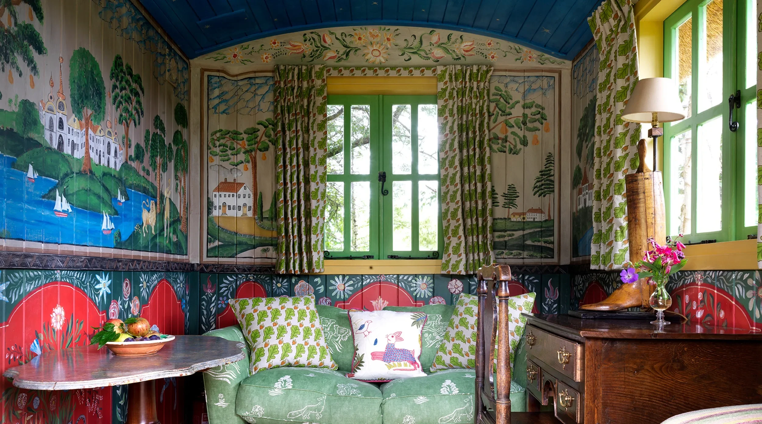 A cozy room with green window frames, floral curtains, and walls decorated with mural paintings of a lakeside village scene. There is a wooden dresser, a green sofa with patterned cushions, a table with a bowl of fruit, and a small lamp. Bright sunlight comes through the windows.