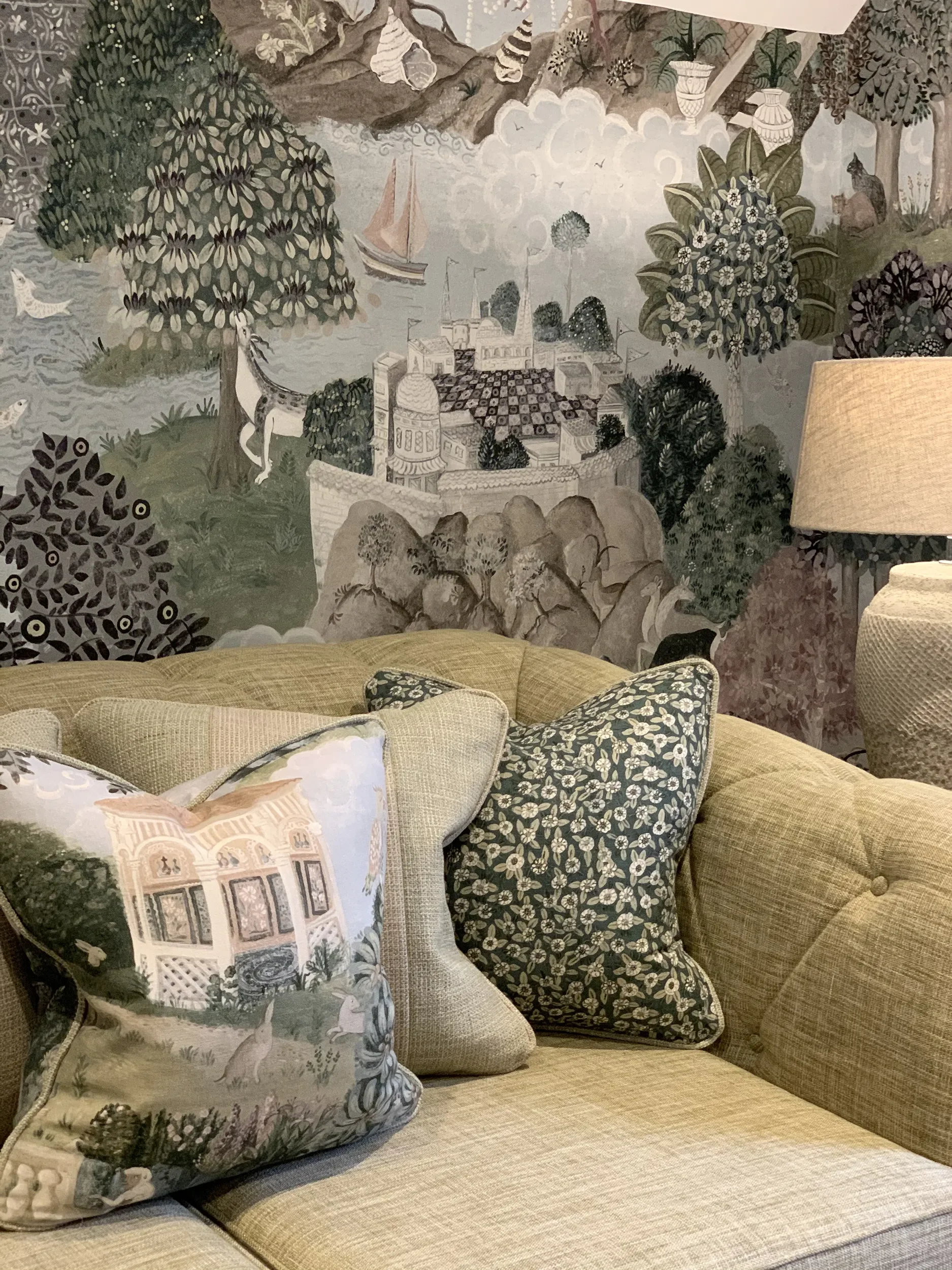 A beige upholstered sofa with a decorative pillow featuring a scenic illustration of a historic building, with lush trees and a garden.