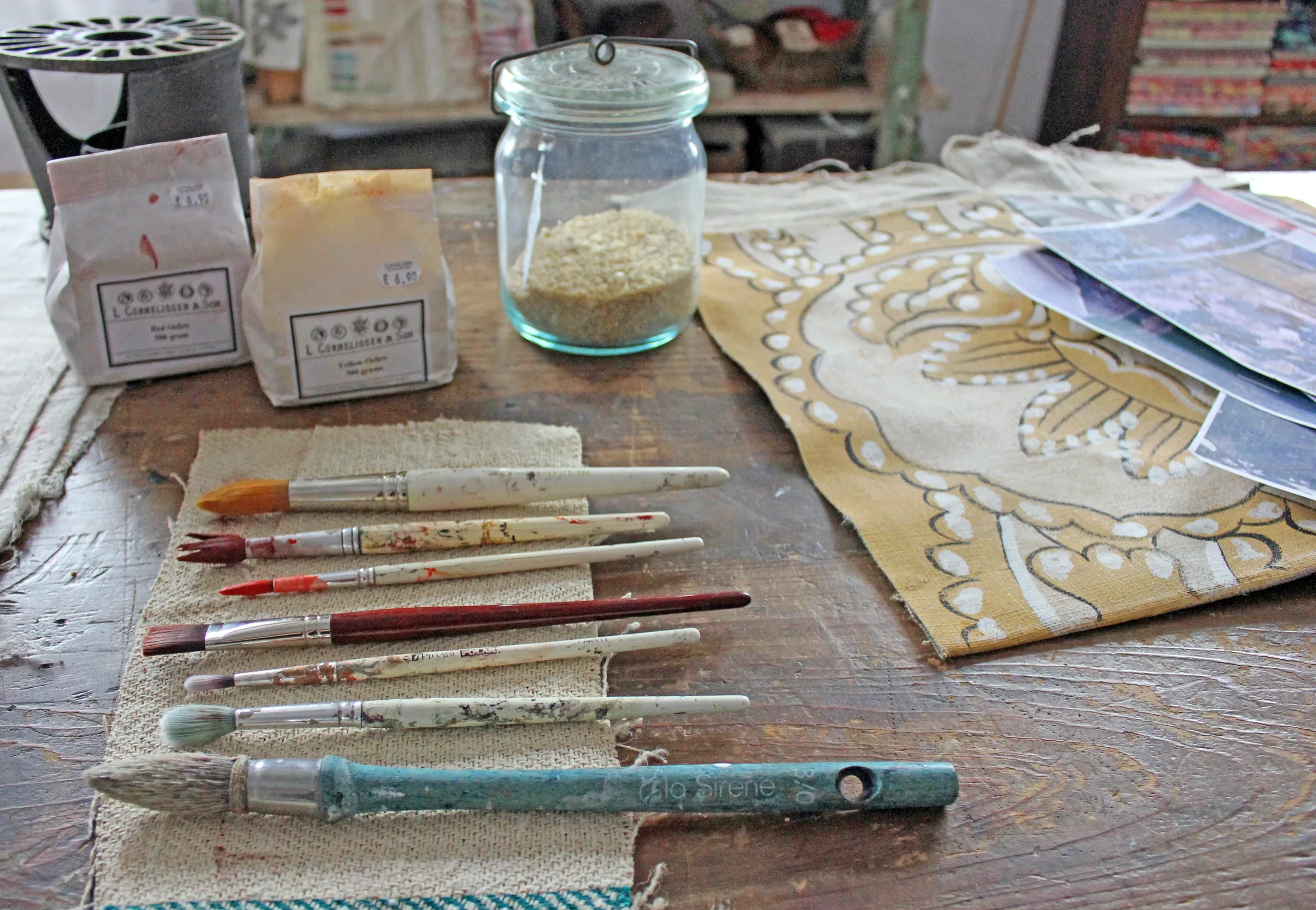 Paintbrushes with various colors of paint resting on a small cloth on a wooden table, with paint supplies, paper, and fabric in the background.