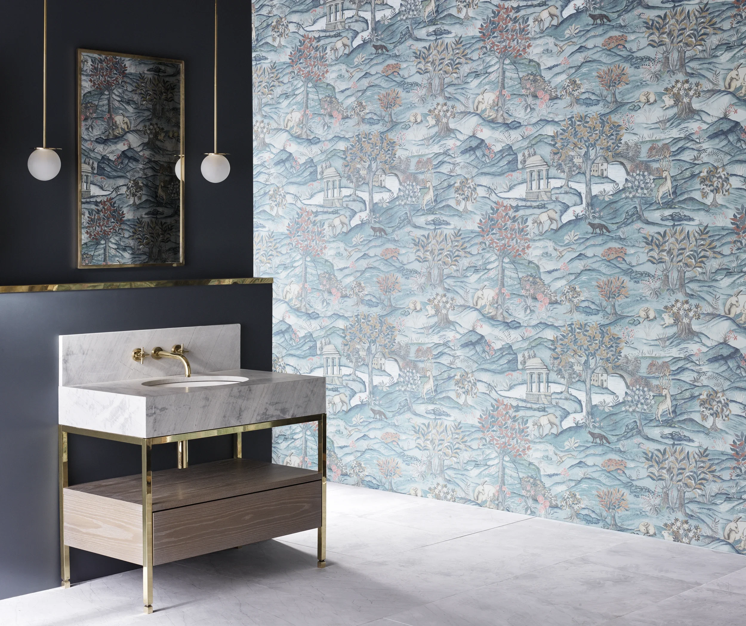 A bathroom with dark blue walls and a large decorative wallpaper featuring a scenic landscape with mountains, trees, animals, and ruins in pastel colors. There is a white marble sink with a gold faucet and a black shelf underneath, along with two modern round pendant lights with white globes hanging from the ceiling.