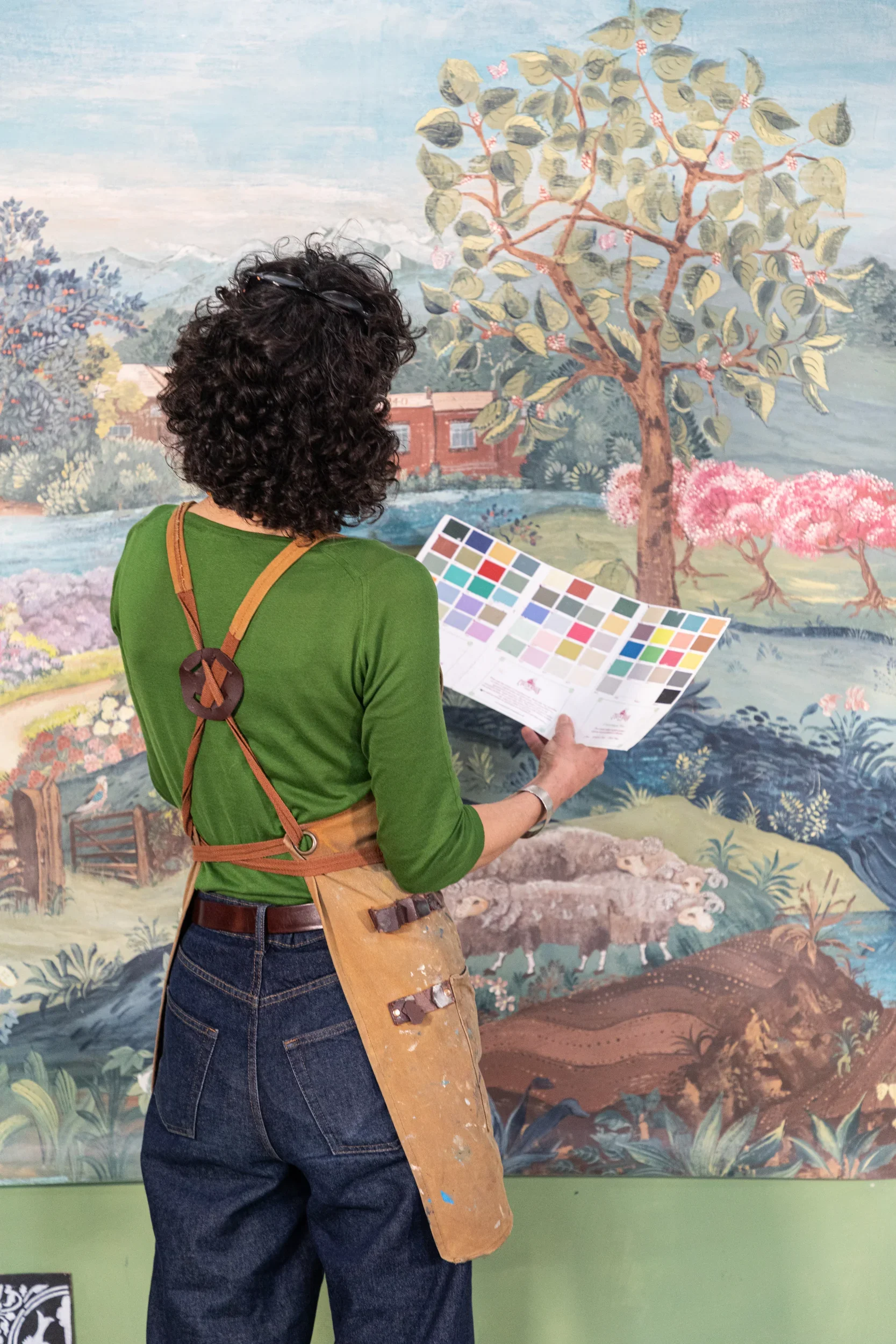 Melissa White is pictured painting the John Smedley 240th anniversary mural. She is wearing an apron with blue jeans and a green jumper. She is back to camera holding a colour swatch.