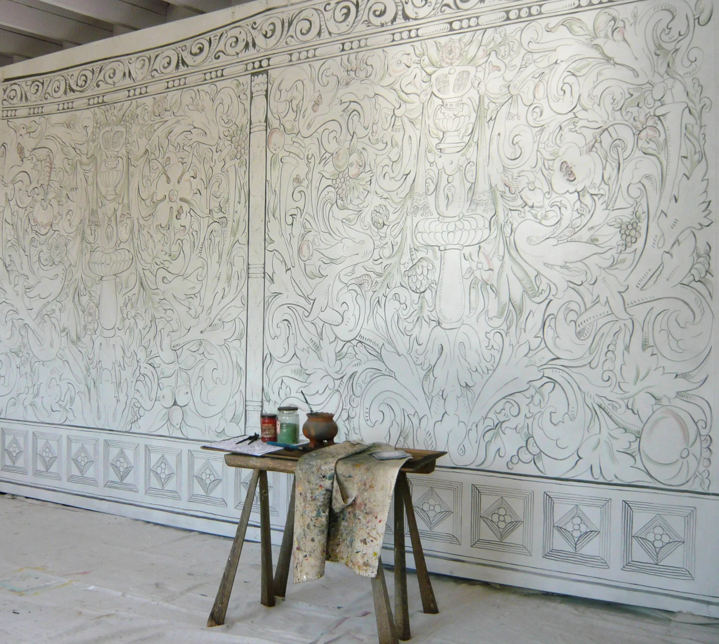 Art supplies, including paint bottles and brushes, are on tables and a stoolin front of Melissa White's Melissa White's largescale multivine painted cloths for Stiffkey Old Hall, Norfolk.