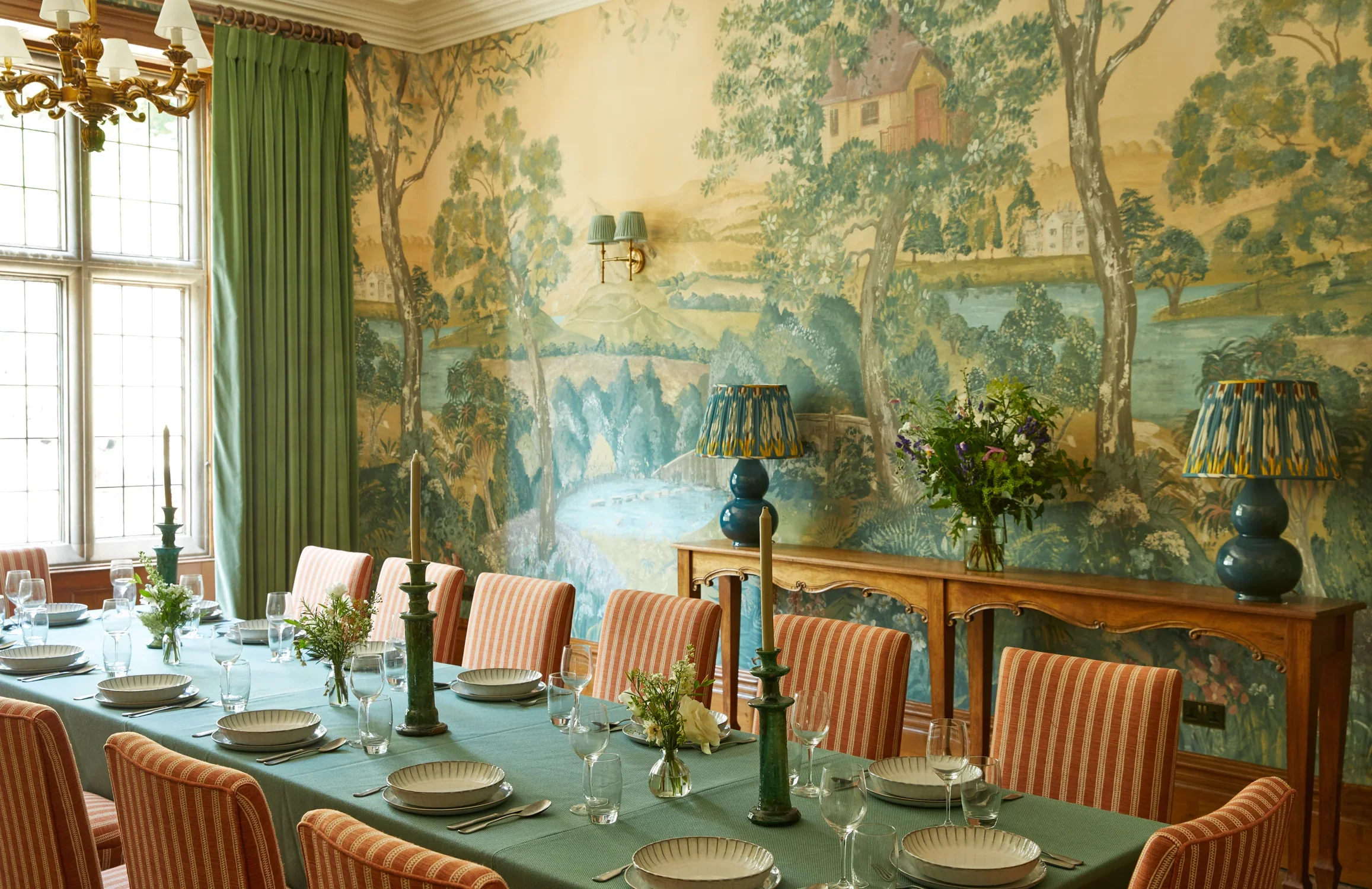 A dining room decorated with a large wall mural depicting a lush landscape with trees, a river, and buildings. The room features a long table set with white dishes, glasses, floral centerpieces, and green tablecloths. The chairs have pinkish-orange striped upholstery. There are green curtains covering a large window, and green lampshades on two table lamps and a wall sconce.