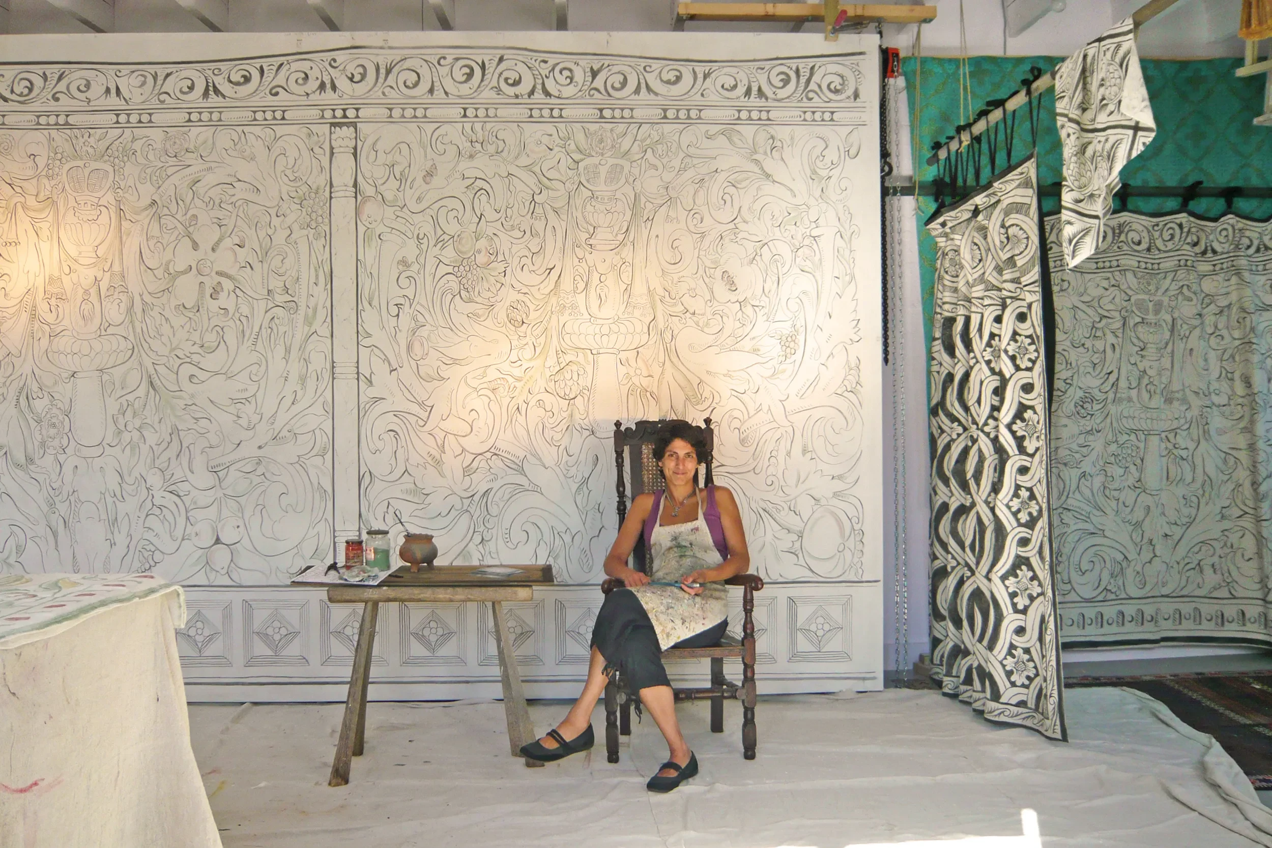 Melissa White sitting on a wooden chair in front of her largescale multivine painted cloths for Stiffkey Old Hall, Norfolk.